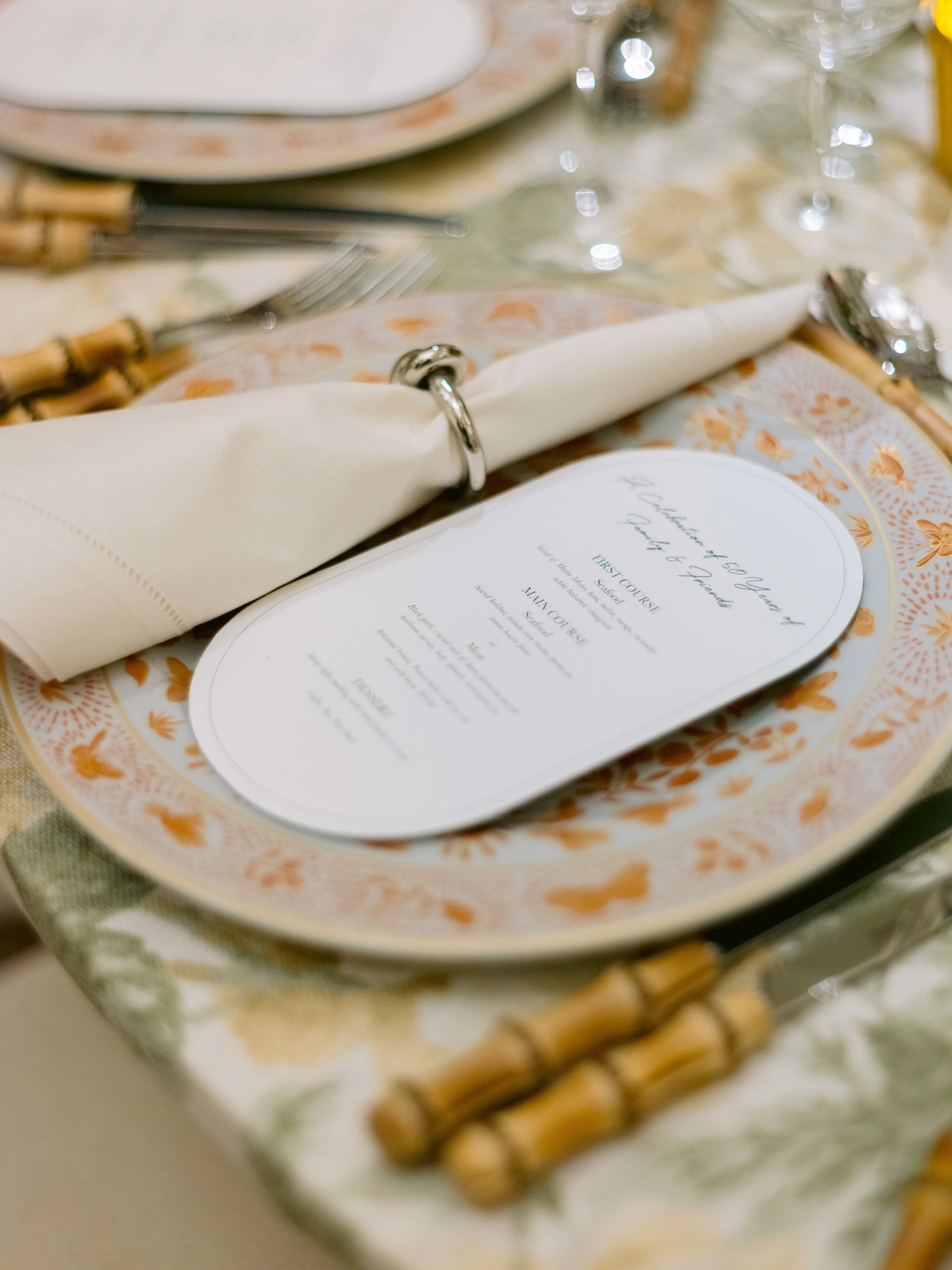 A table setting with a white cloth napkin secured with a silver ring, a menu, a plate, cutlery, and a decorative placemat with orange and blue patterns.