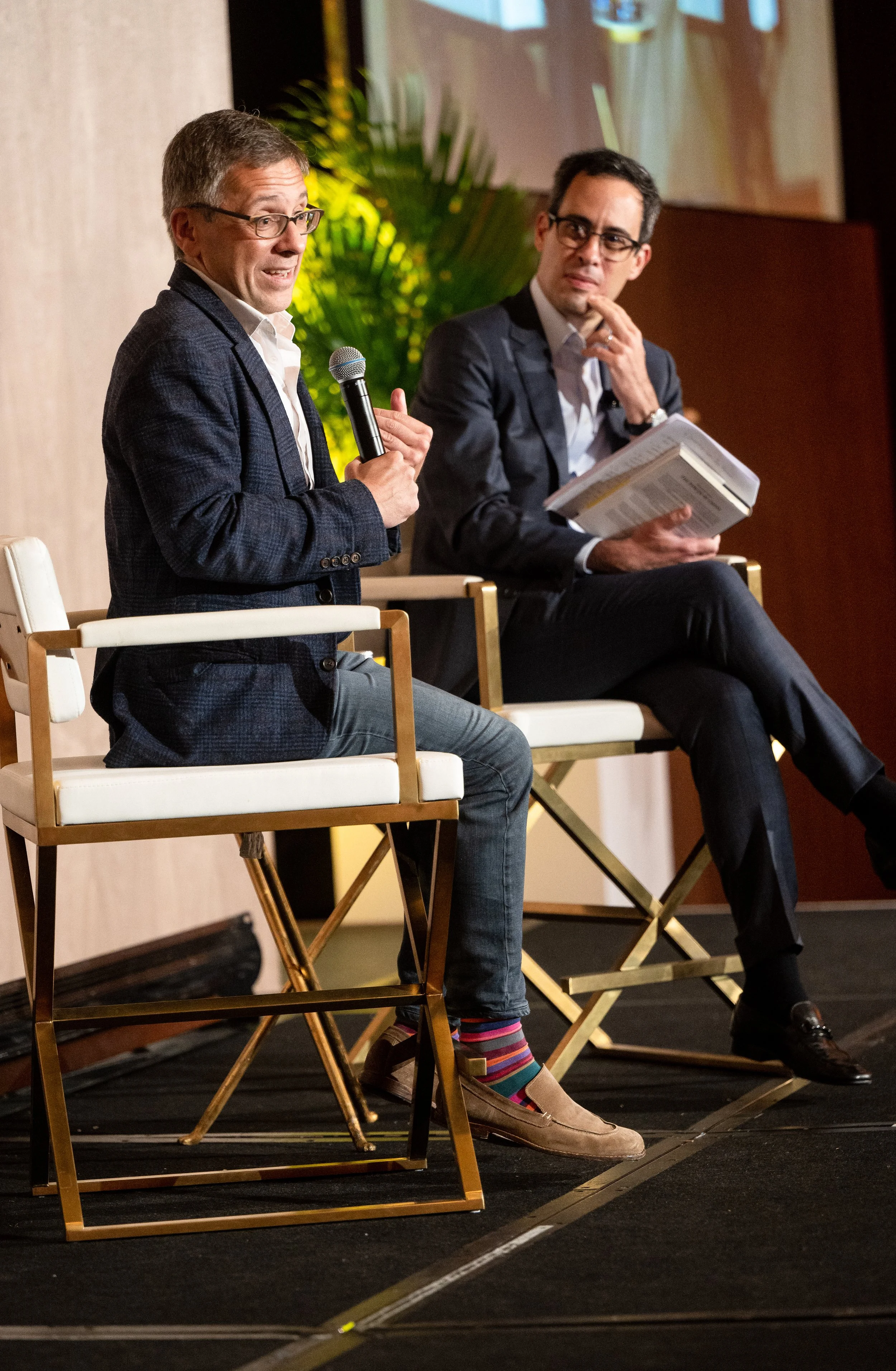 Two men sitting on a stage having a discussion or panel event. One man is speaking into a microphone, the other is holding a book and listening. Both are dressed in suits.
