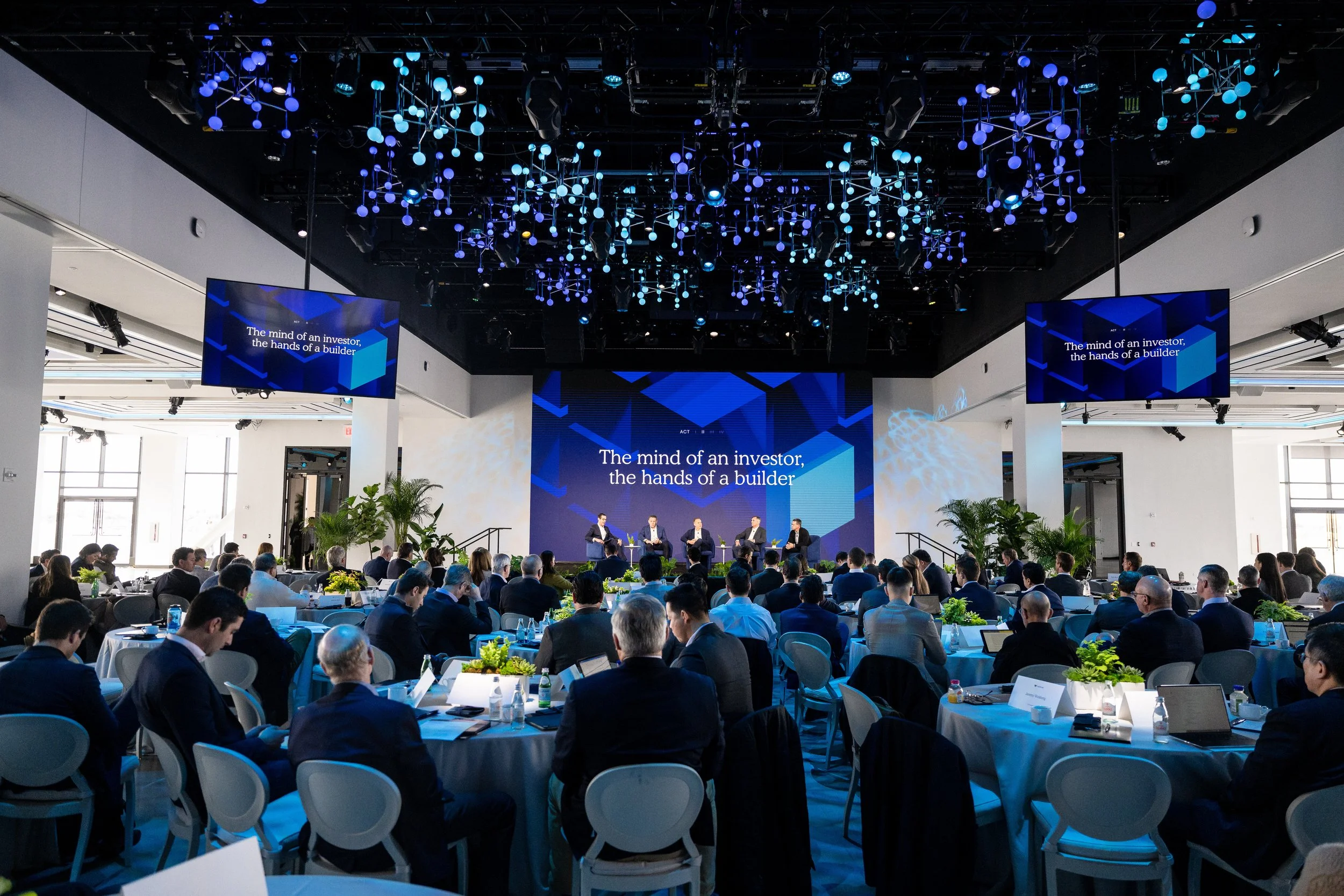 A large conference room filled with people attending a panel discussion. There are multiple screens displaying the phrase "The mind of an investor, the hands of a builder." The ceiling has decorative blue and white hanging lights, and there are plant