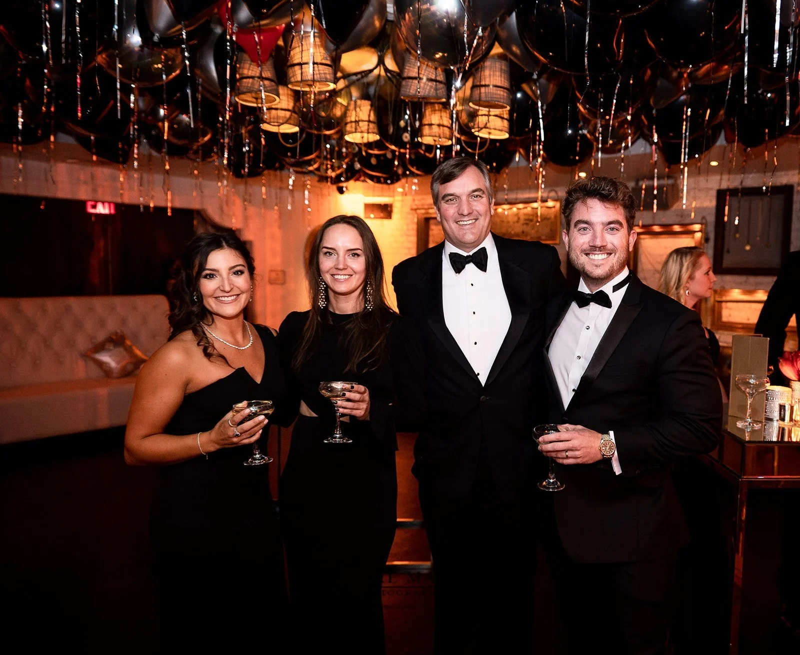 Four people dressed in formal evening attire at a celebration or event, smiling and holding drinks in a dimly lit venue with decorative black balloons and string lights above.