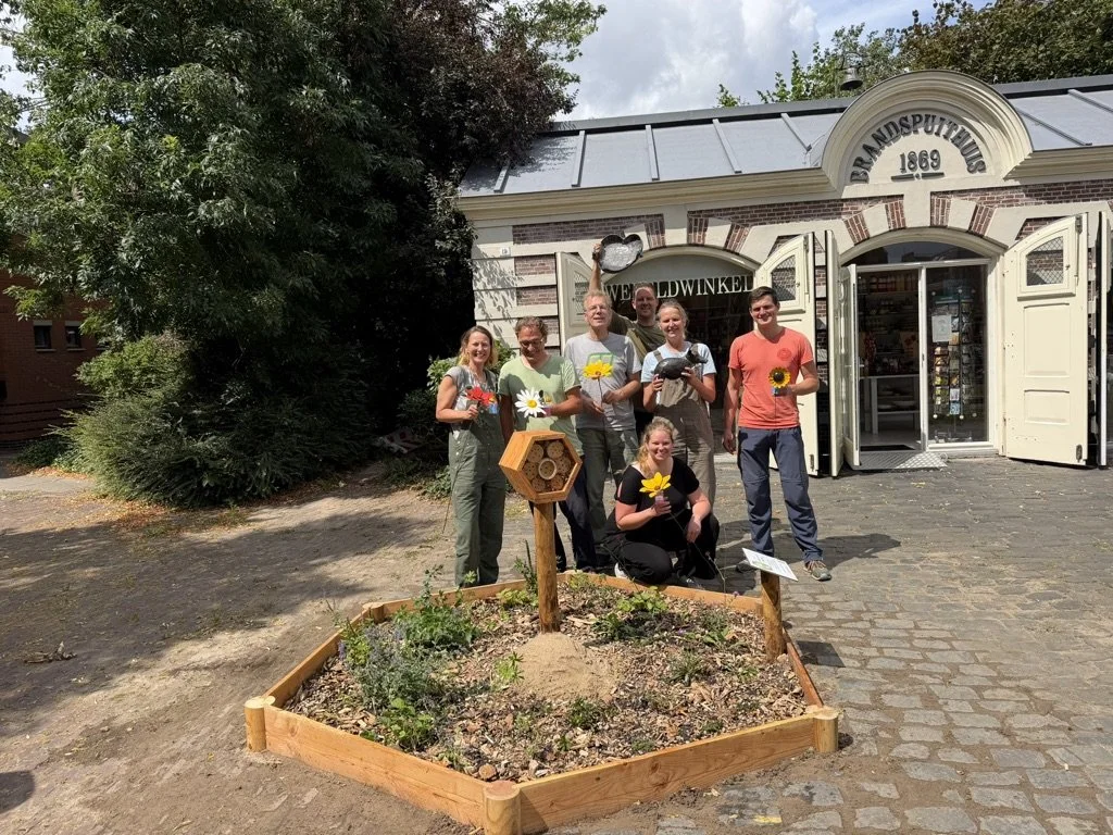 Group of seven people standing outside a historic building with open doors, holding flowers, and posing for a photo. The building has a sign that reads "Handspuitmuseum 1869." In front of the group is a small garden bed with plants and a wooden beehive decoration, and a sign next to it.