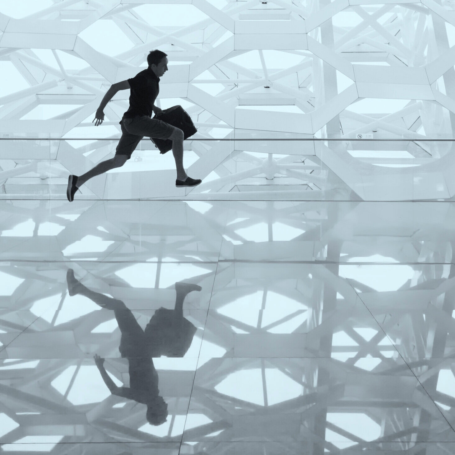 A man carrying a bag is running across a glossy, reflective floor inside a modern building with a geometric, white architectural ceiling.