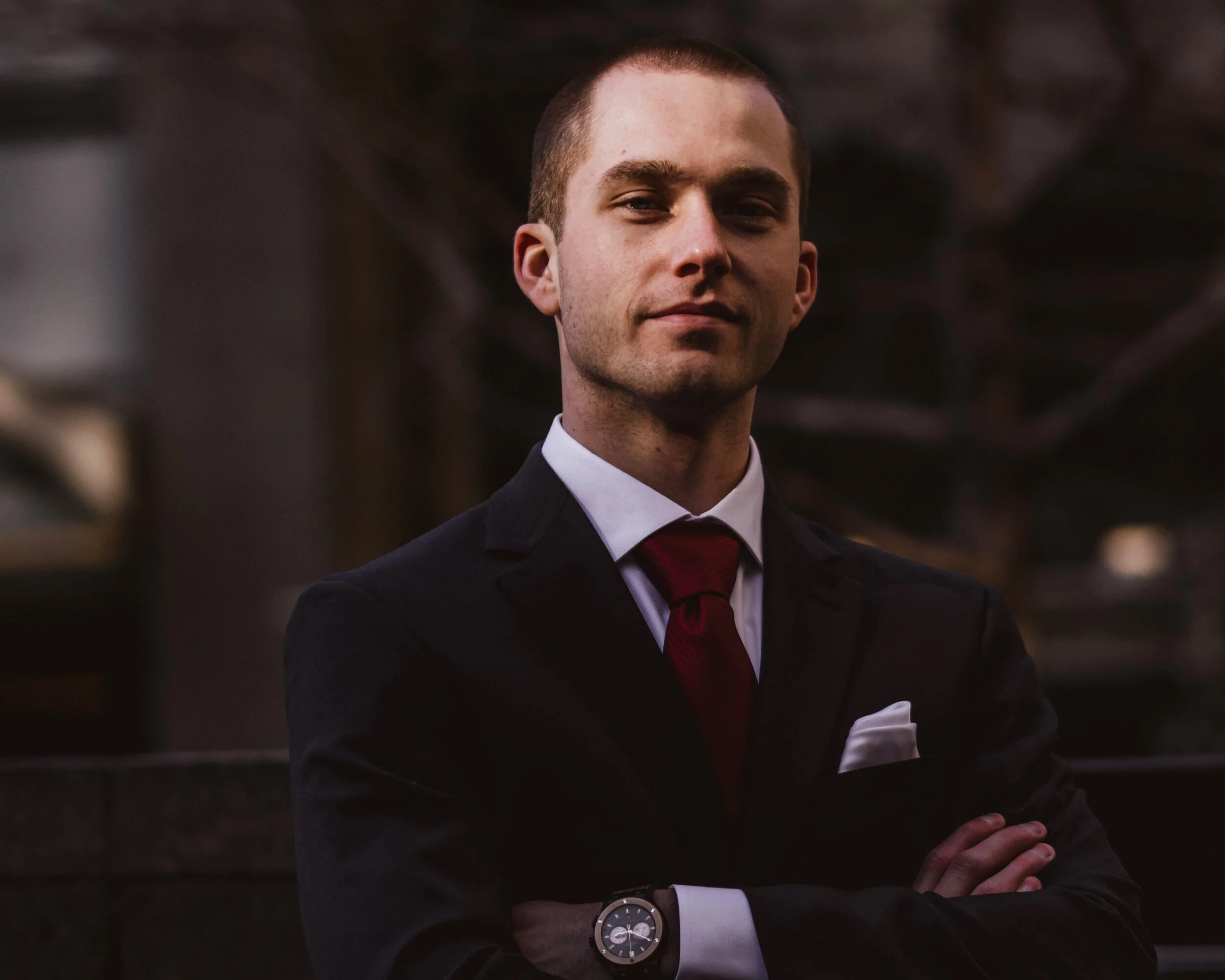 A confident young professional man in a dark suit with a white shirt, red tie, and a wristwatch, standing with arms crossed in an indoor setting.