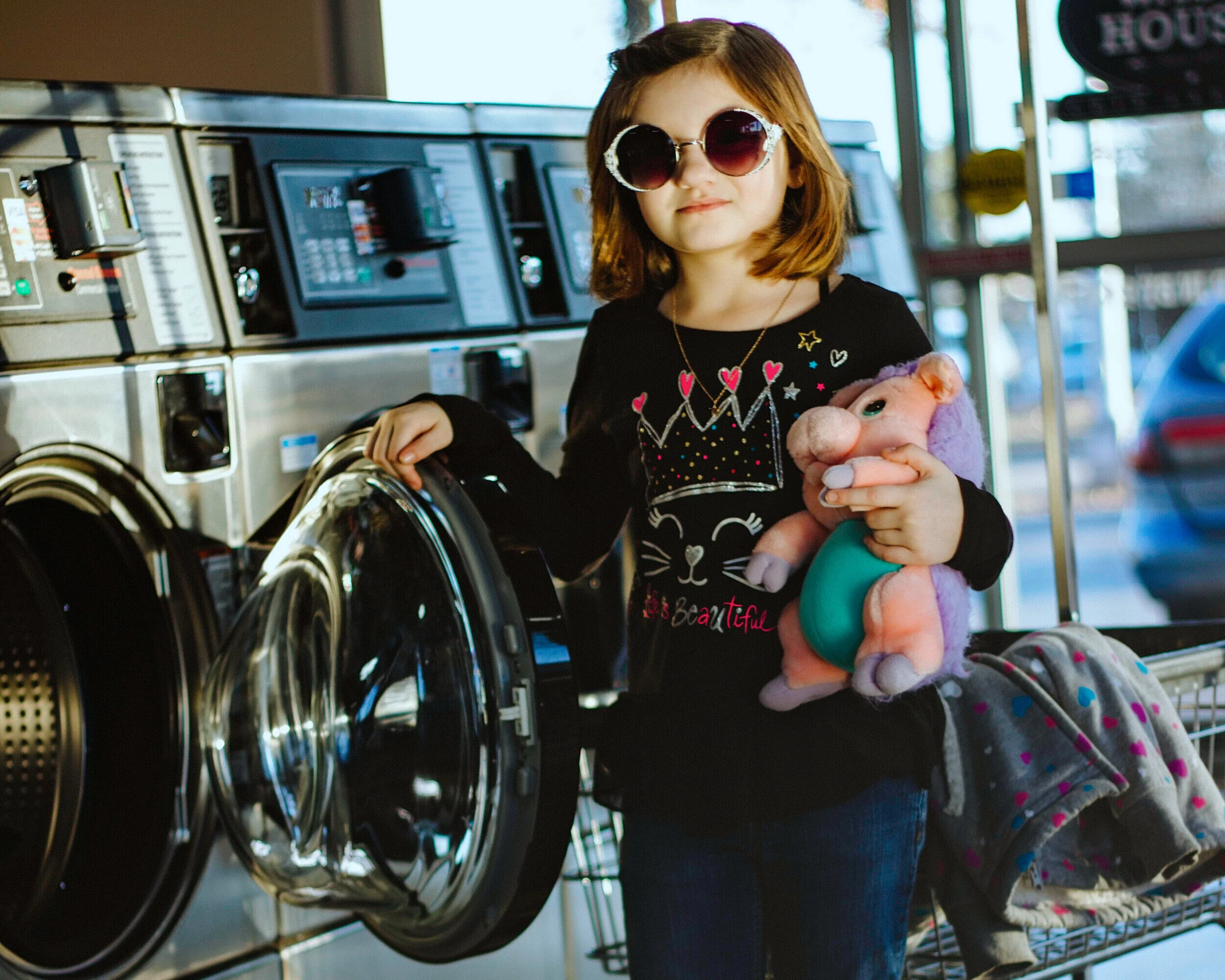 A young girl with shoulder-length red hair, wearing sunglasses and a black shirt with a cat face and crown design, is standing in front of a row of washing machines at a laundromat. She is holding a plush pig toy and has a shopping cart with clothes and a blanket next to her.