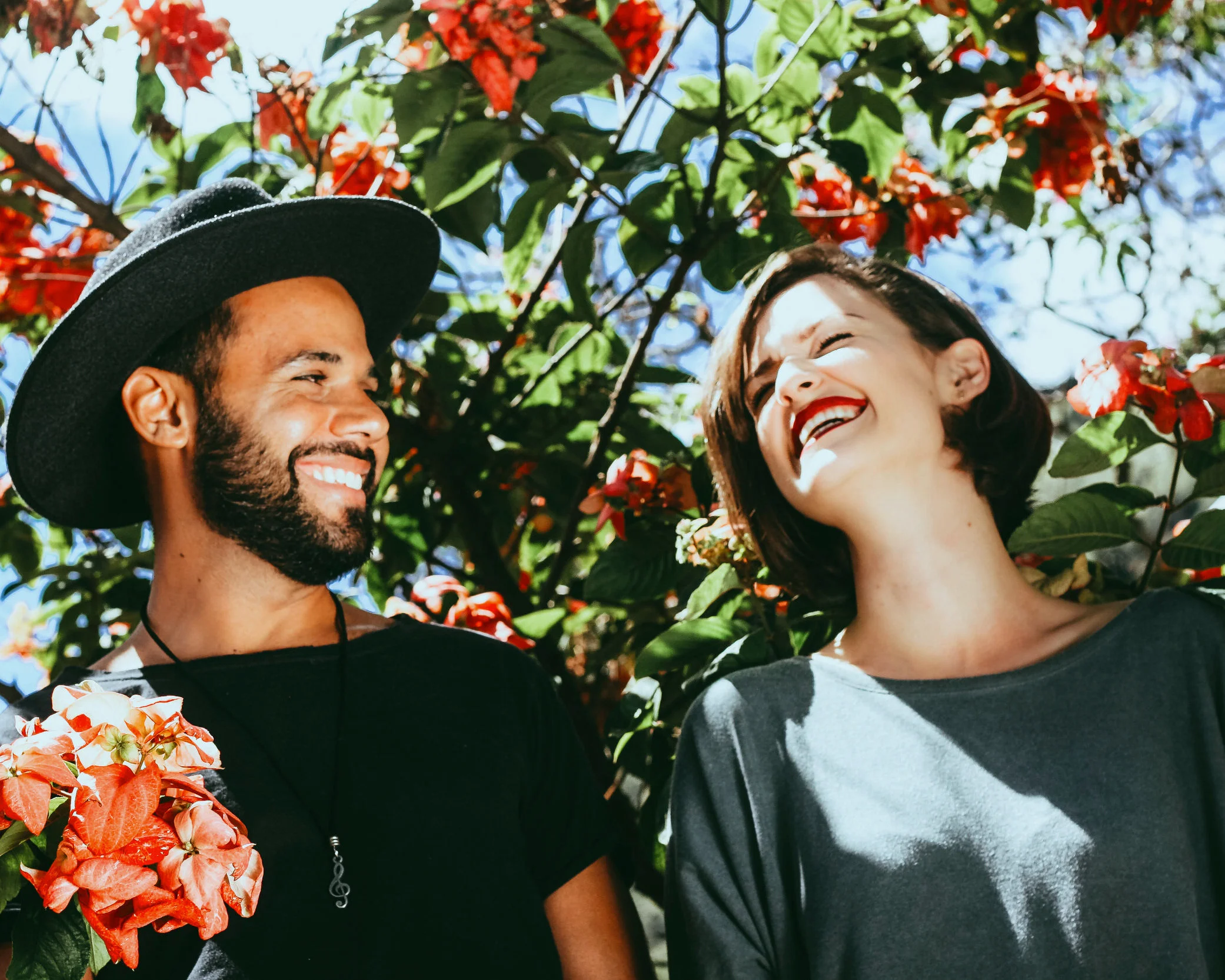A BIPOC man and woman smiling and laughing outdoors among red flowers and green leaves on a sunny day.