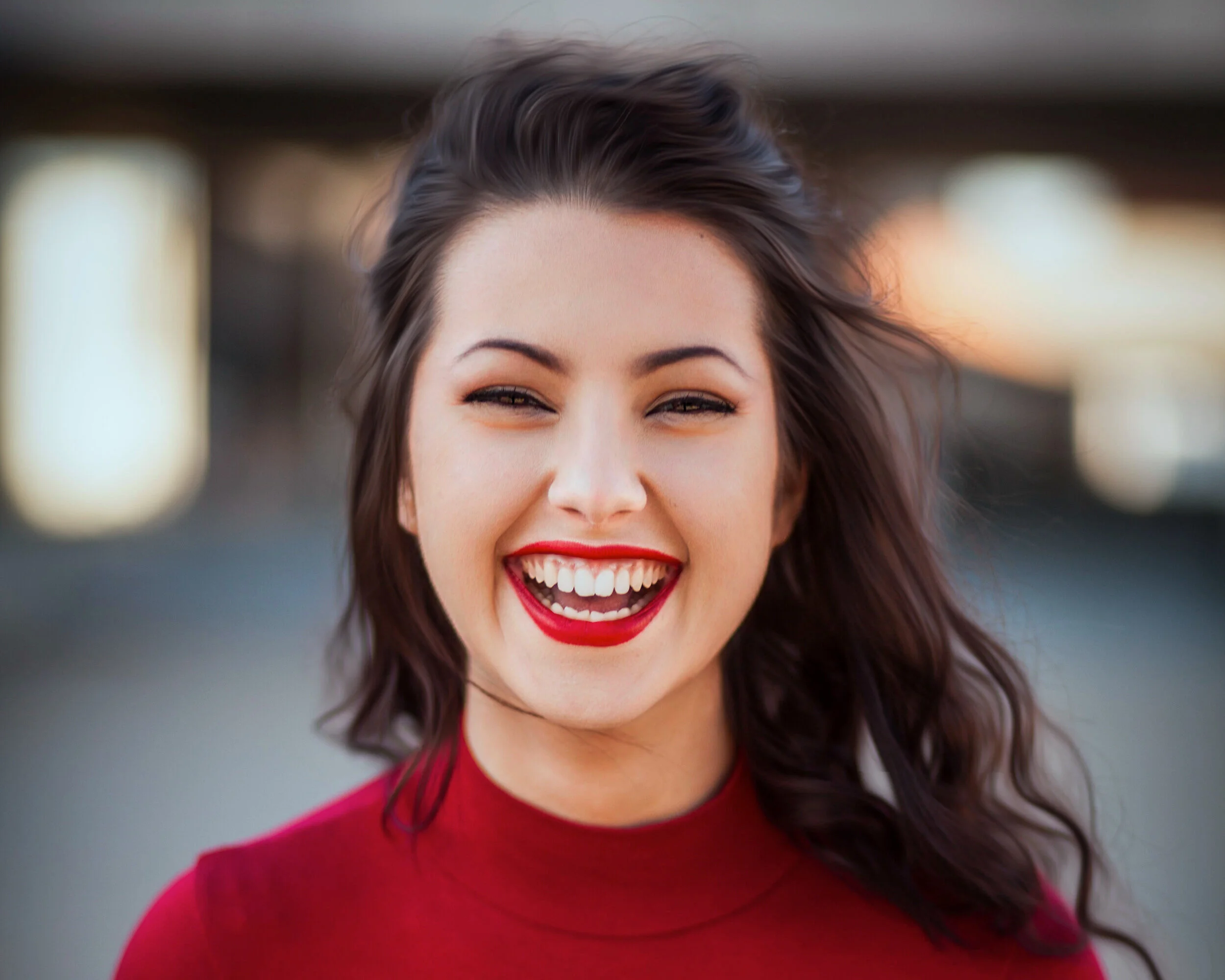 A young Latinx woman with dark wavy hair smiling broadly, showing her teeth, wearing red lipstick and a red top, outdoors during sunset.