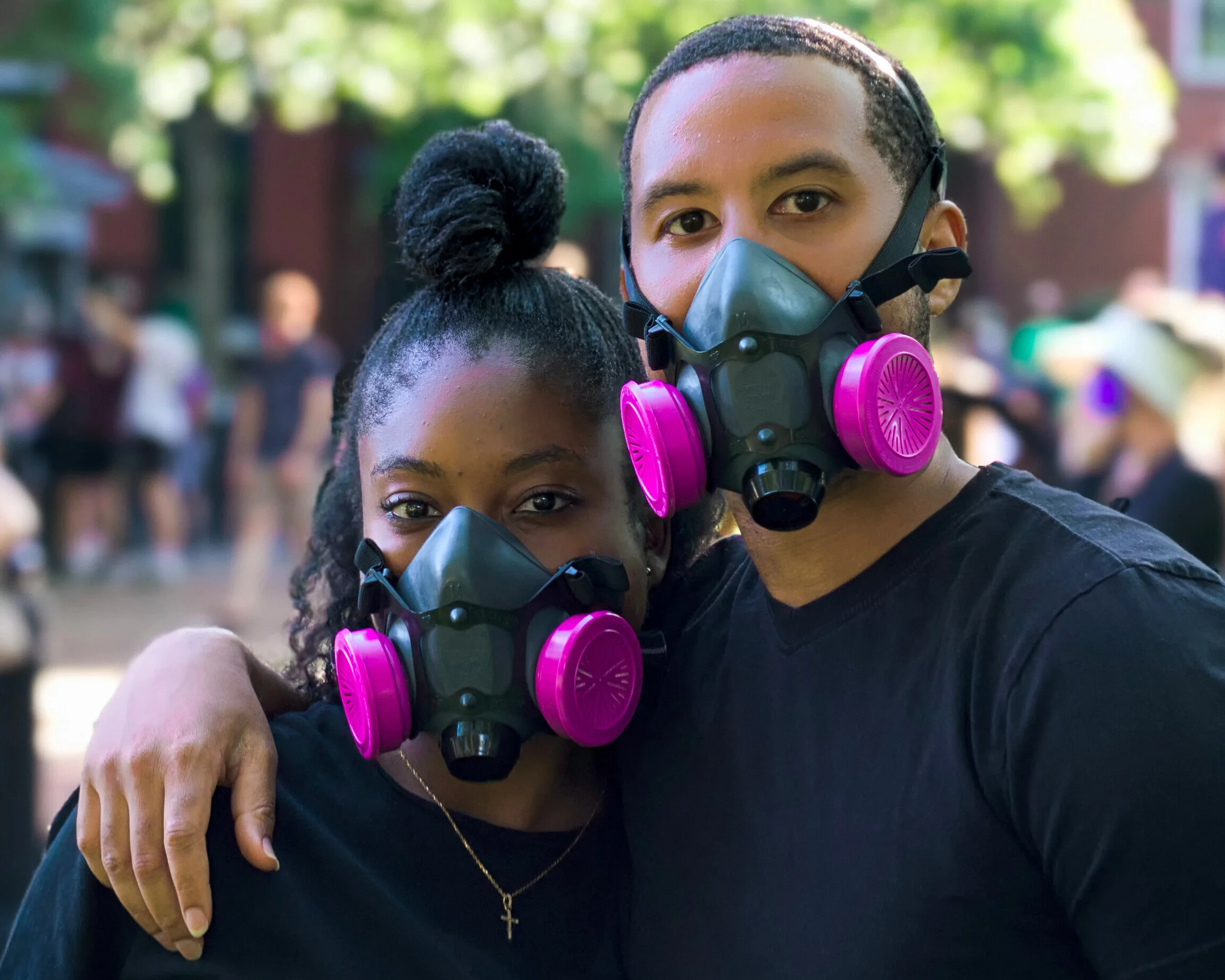 A BIPOC man and woman wearing black masks with pink filters, standing close together outdoors, with other people in the background.