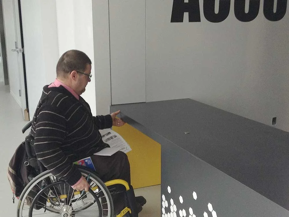 Utilisateur de fauteuil roulant à l'accueil (comptoir évidé pour s'approcher) du centre communautaire Lebourgneuf. 