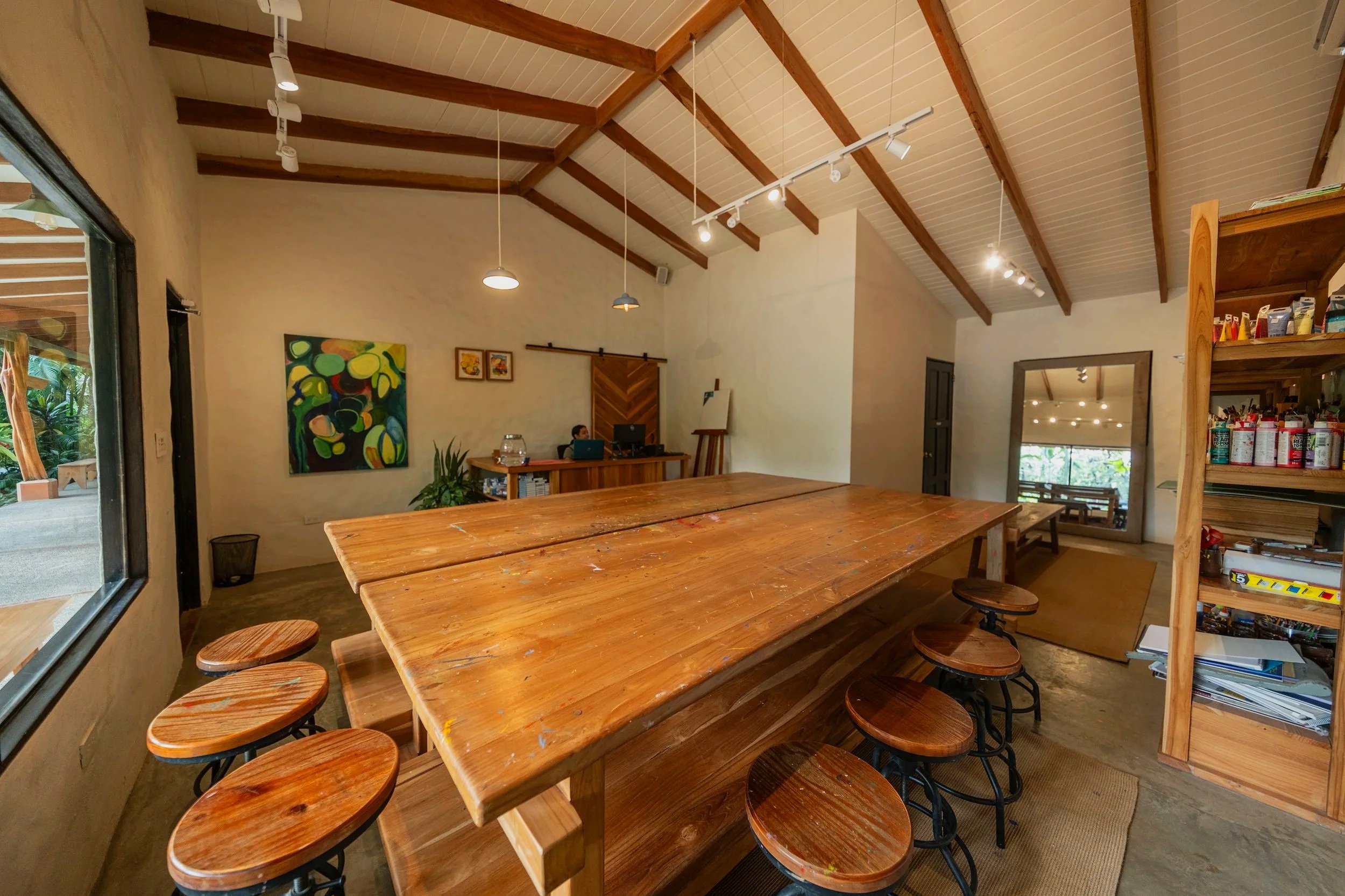 Interior of a rustic room with wooden beams on the ceiling, a large wooden table, round wooden stools with metal legs, and shelves filled with various items. A person is walking by, creating a motion blur effect.