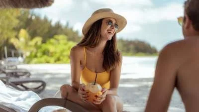 A woman wearing a yellow swimsuit and sun hat sitting on the beach, holding a coconut drink with a straw, talking to a man sitting nearby.