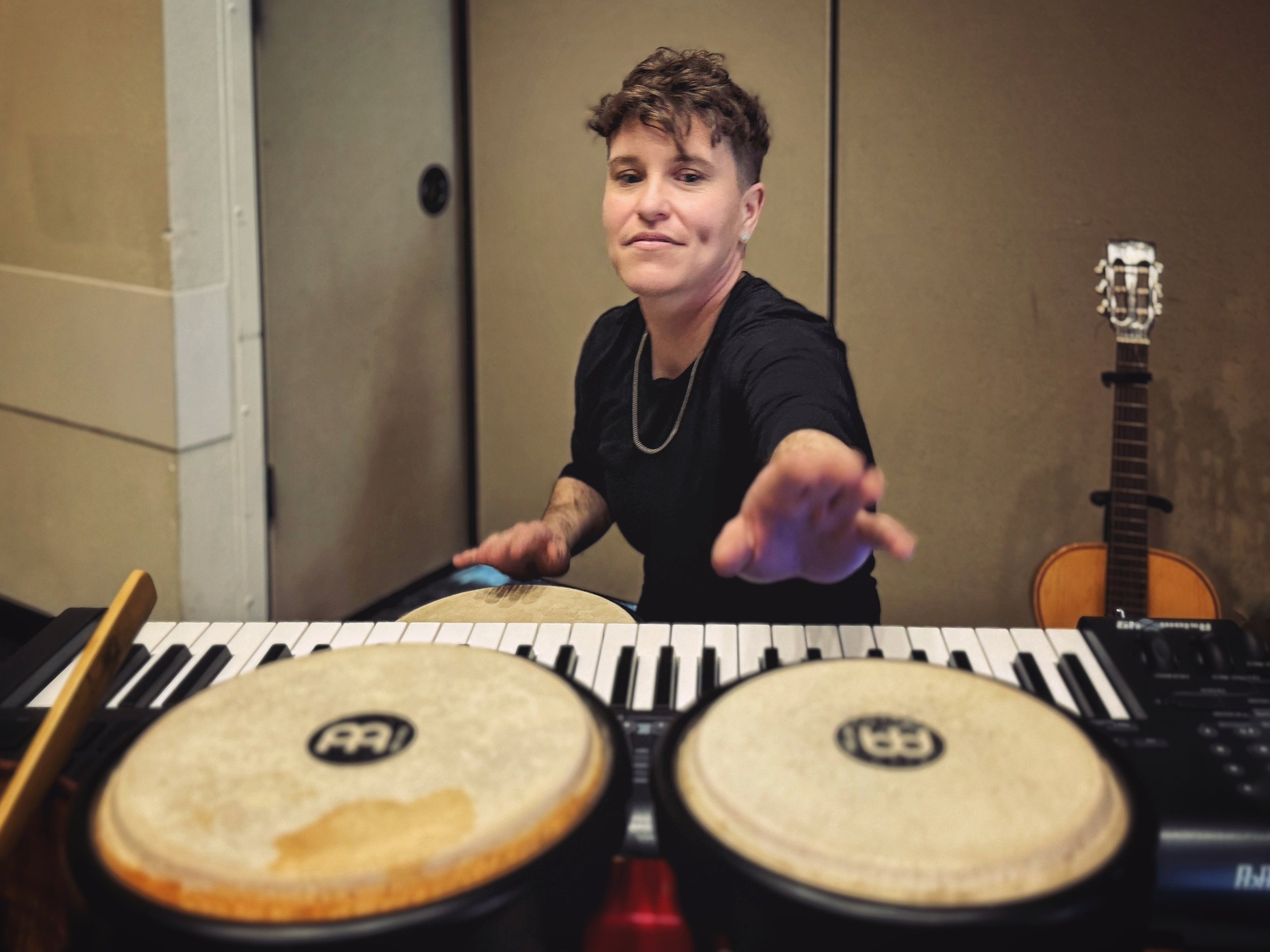 A musician is smiling while seated at a keyboard, surrounded by drums and a guitar