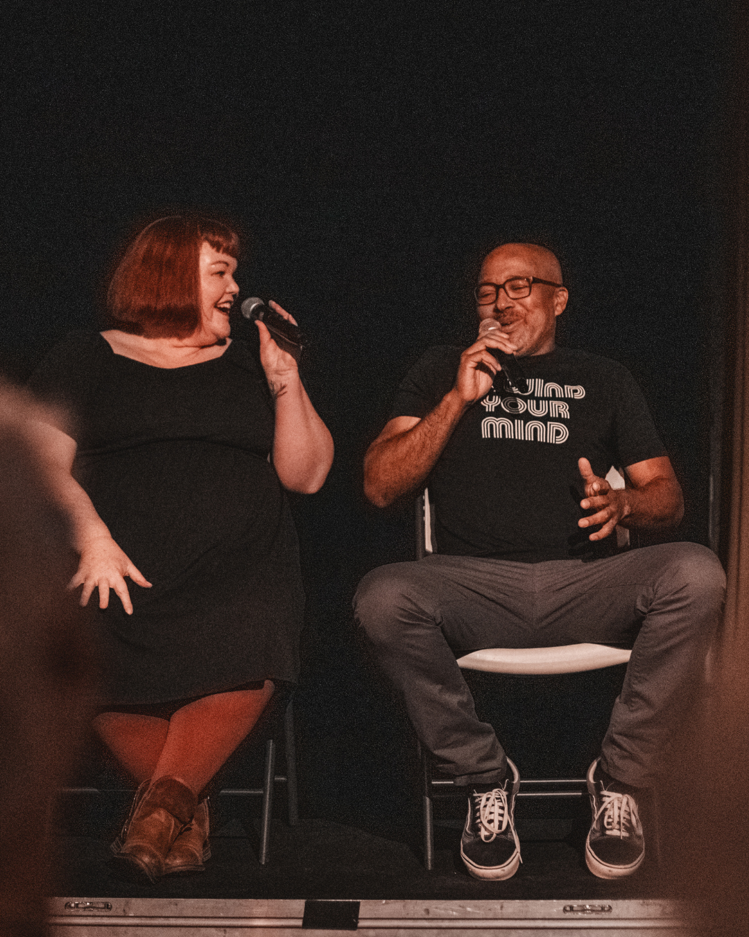 A woman and a man sit side by side on stage, both with microphones. She interviews the man while he tells his story.