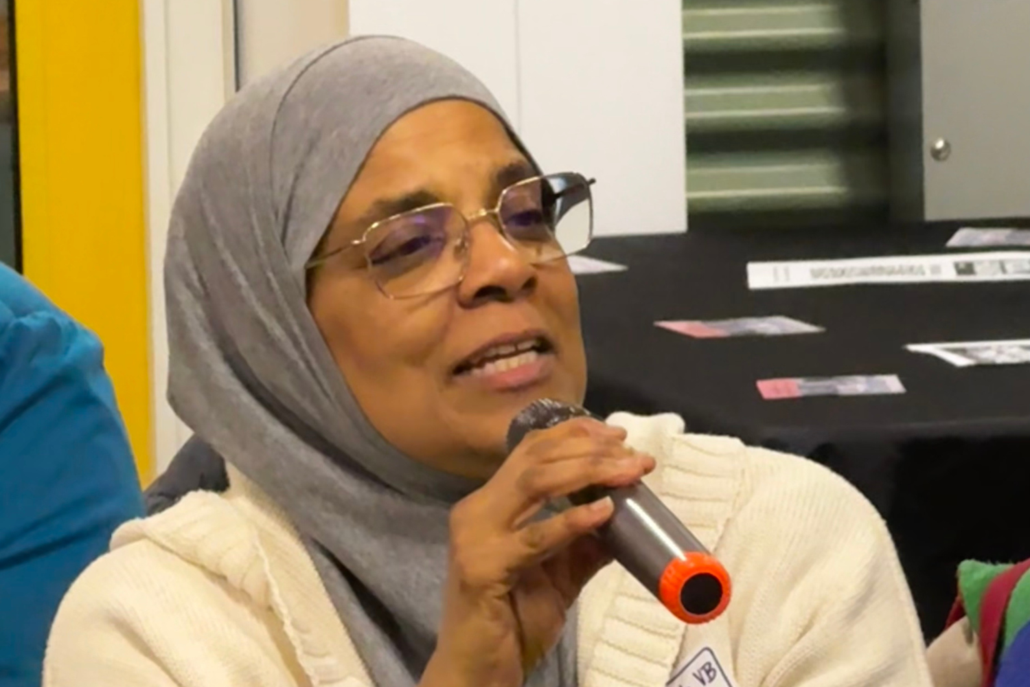 A woman wearing glasses and a hijab is speaking into a microphone