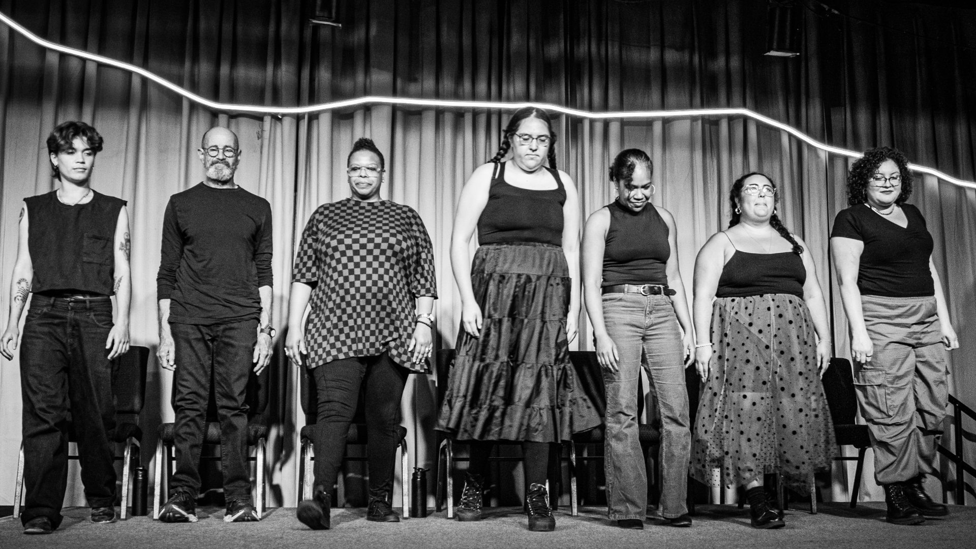 7 actors lined up together, shoulder to shoulder, on stage