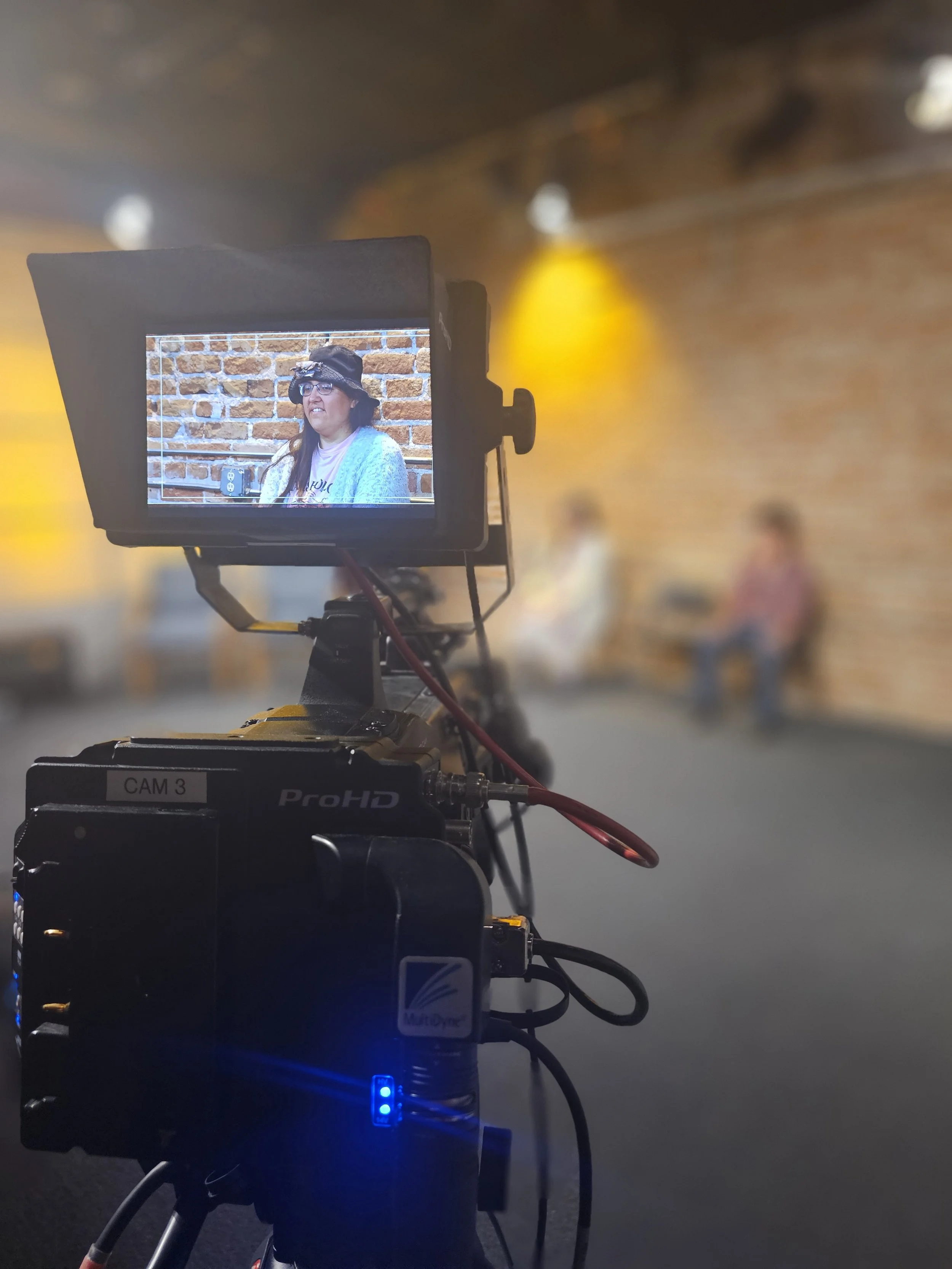 a video camera in a TV studio shows a shot of a human with a hat and glasses smiling