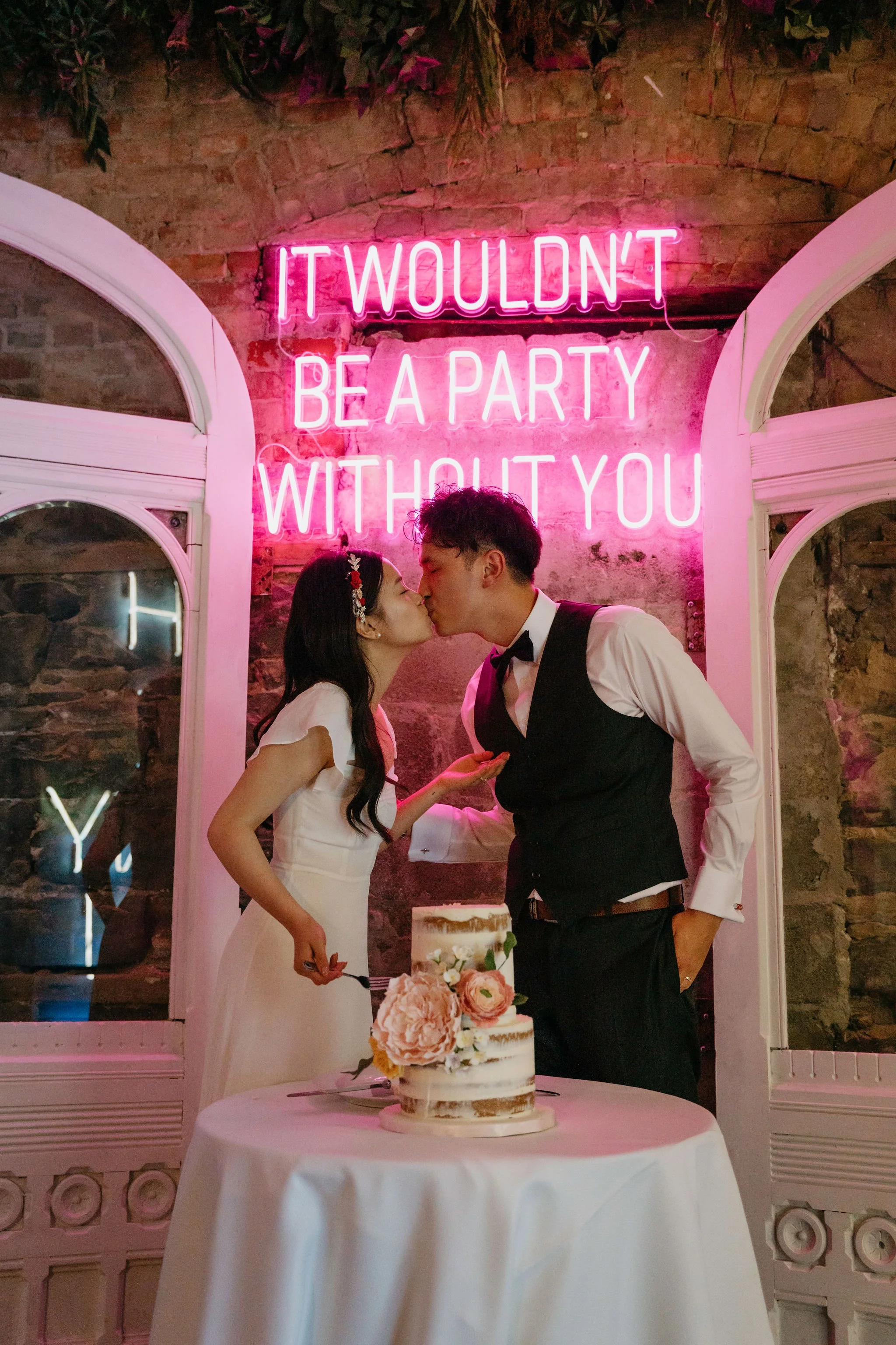 Bride and Groom kissing while cutting cake with pink neon sign behind them