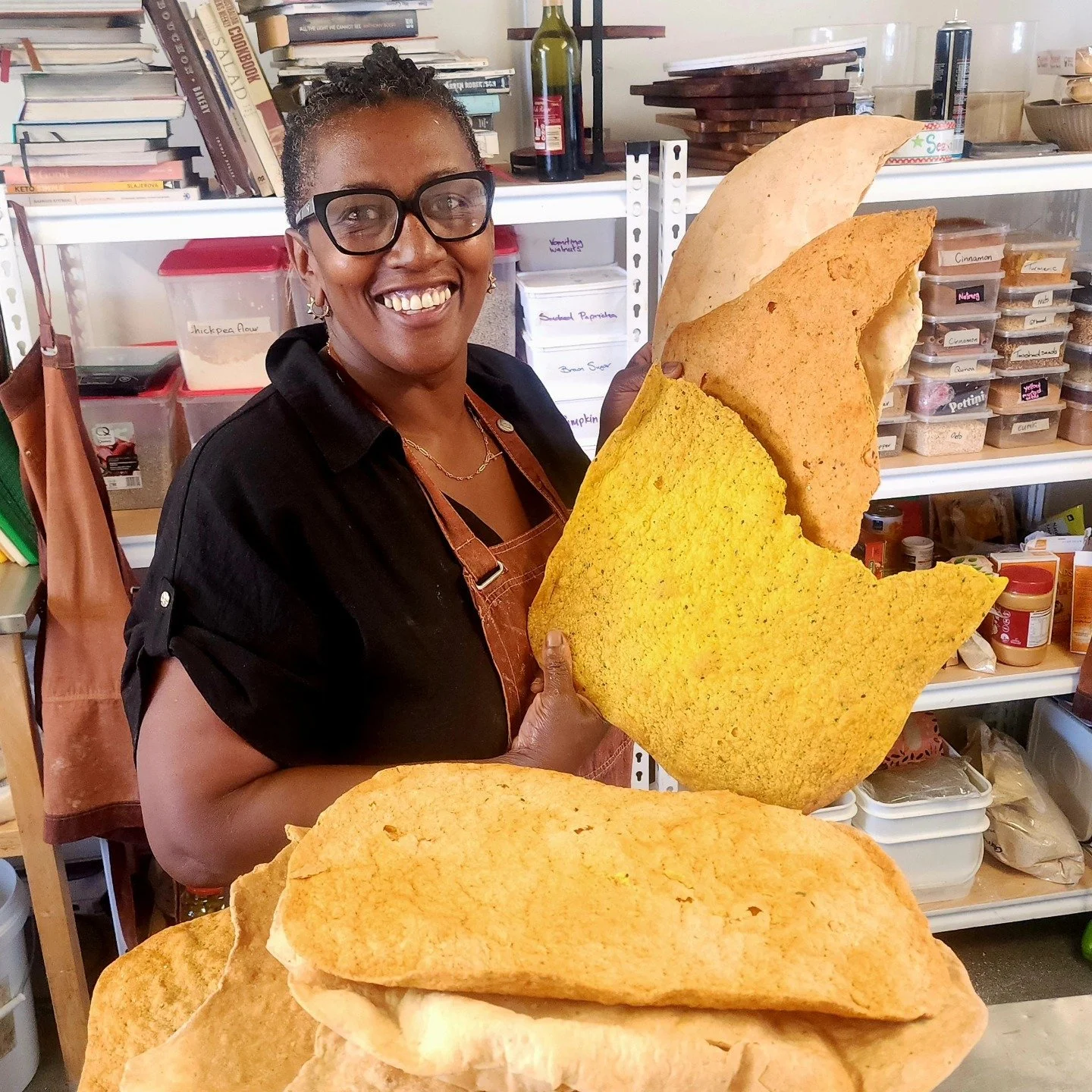 A smiling woman wearing glasses and a black shirt, holding large pieces of bright yellow and orange snack chips in a kitchen or pantry with shelves of food and spices in the background.