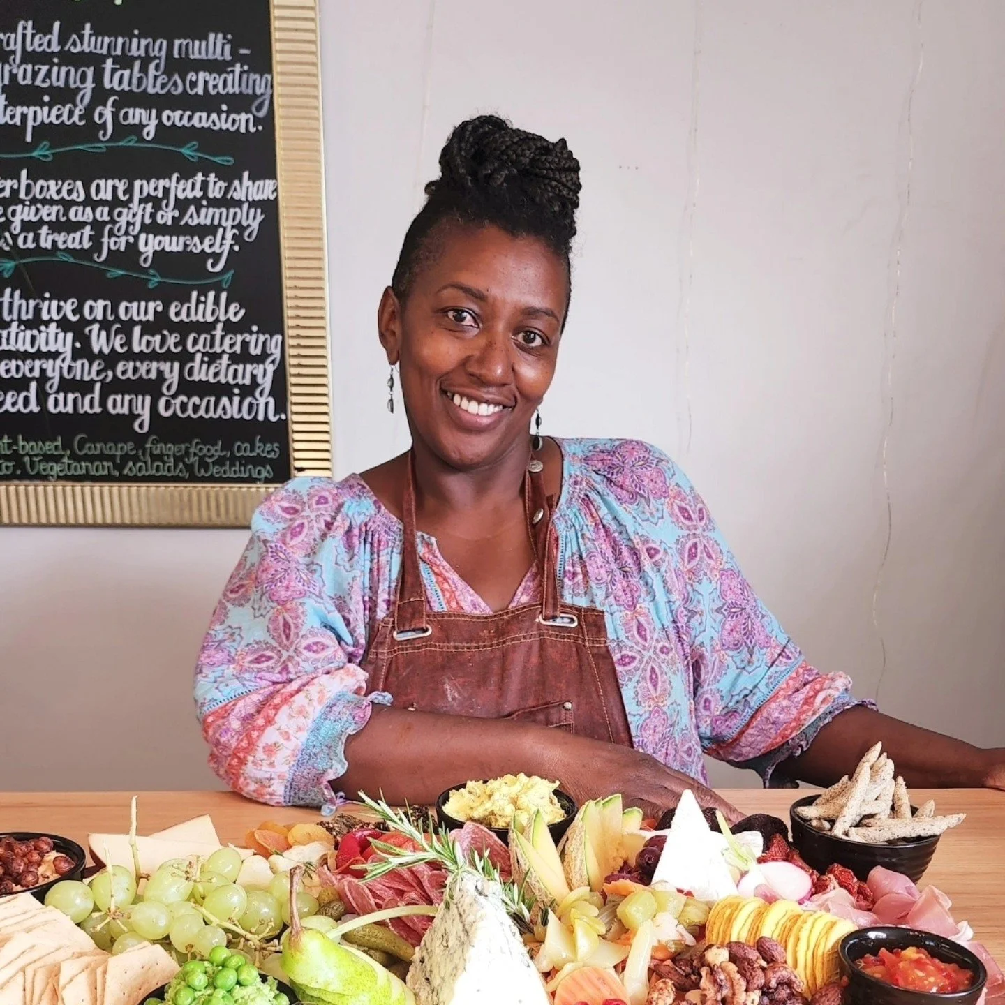 A smiling woman wearing a colorful blouse and a brown apron sitting at a table with a cheese and fruit platter, including grapes, pears, cheese, crackers, and other snacks, in a cafe or restaurant setting.