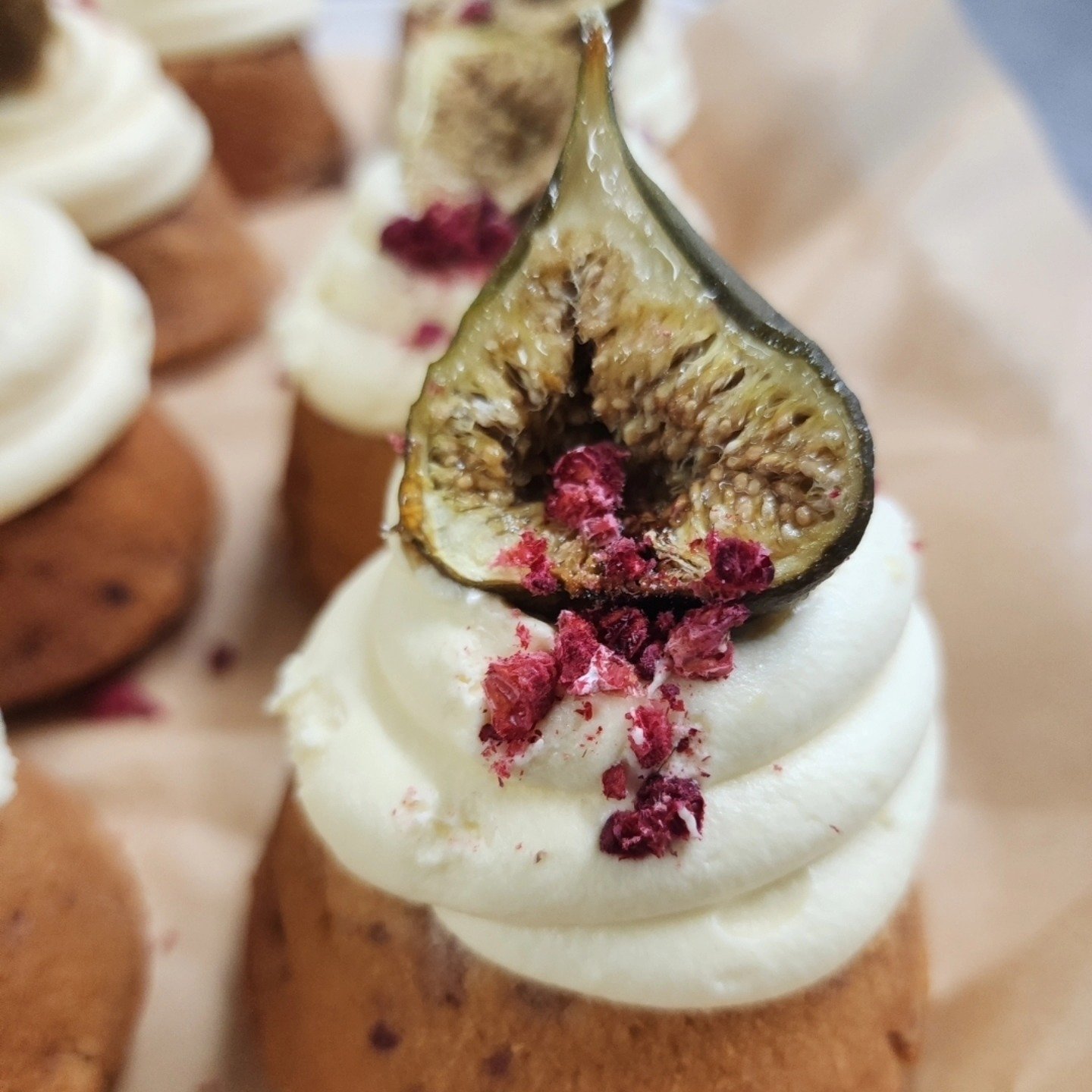 Close-up of a dessert topped with whipped cream, a halved fig, and dried red berries, with a blurred background of similar desserts.