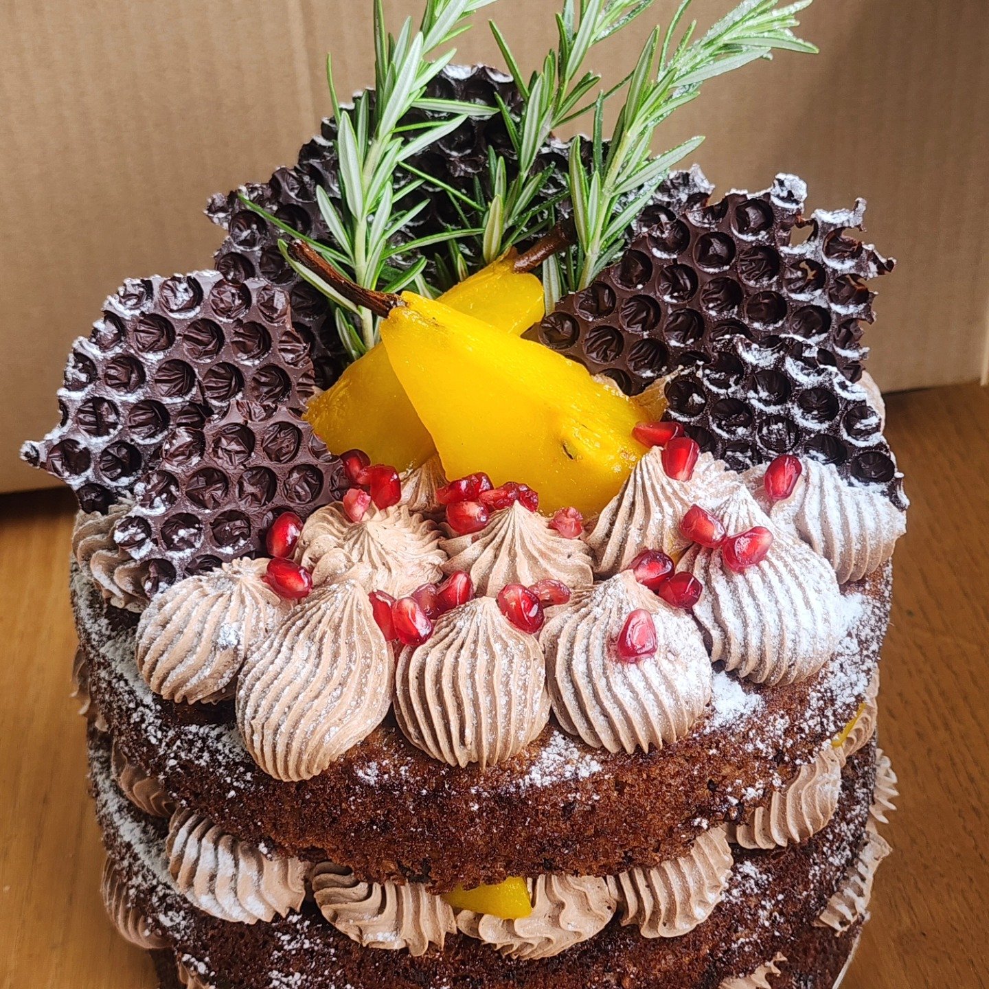A layered chocolate cake decorated with piped chocolate frosting rosettes, red pomegranate seeds, slices of yellow mango, sprigs of rosemary, and chocolate-coated honeycomb pieces, dusted with powdered sugar.