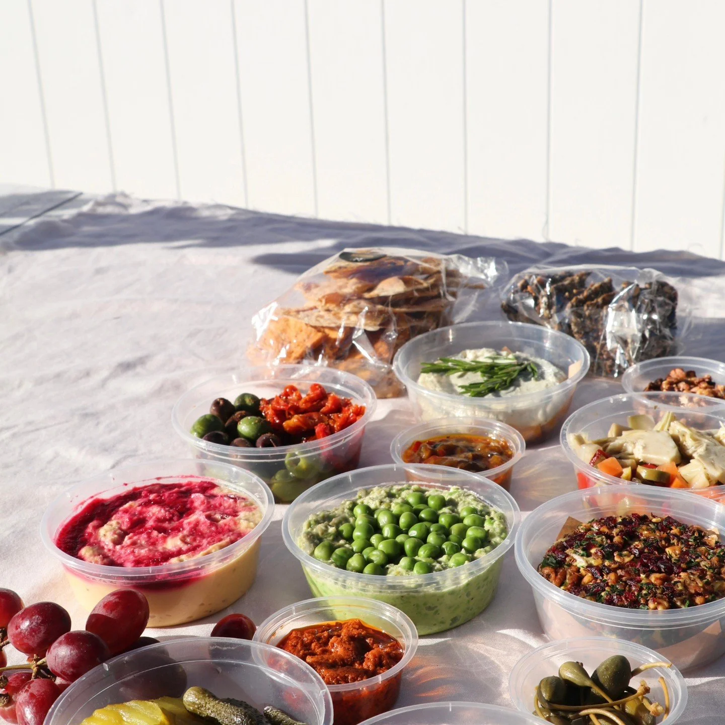 Assorted food items in clear plastic containers, including dips, spreads, olives, grapes, and crackers, with some packaged snacks in the background, laid out on a white table outdoors.