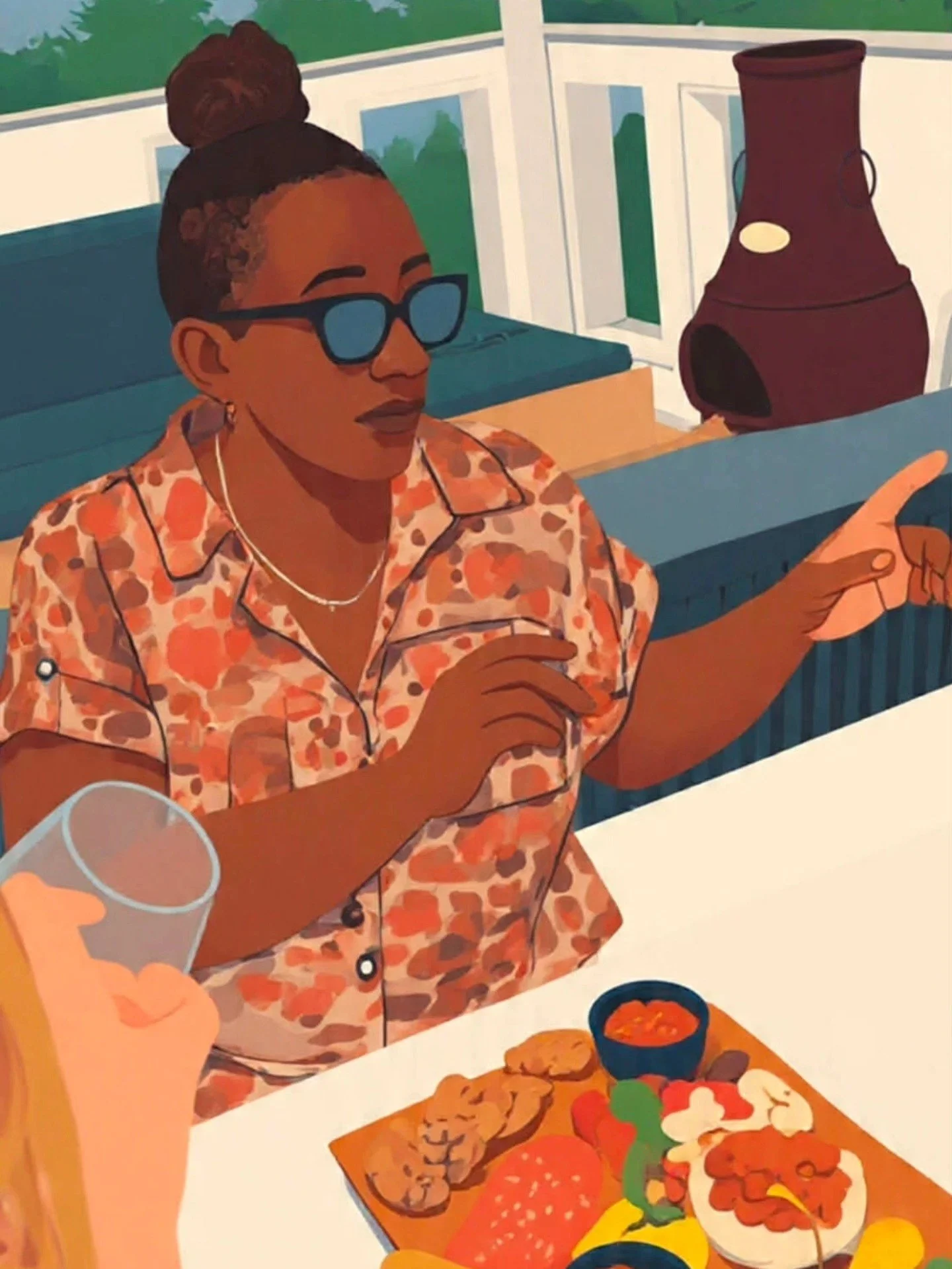 A woman with dark hair in a bun, wearing sunglasses and a floral shirt, sitting at a table with a variety of food, including grilled items, vegetables, and dips, during a meal or gathering.