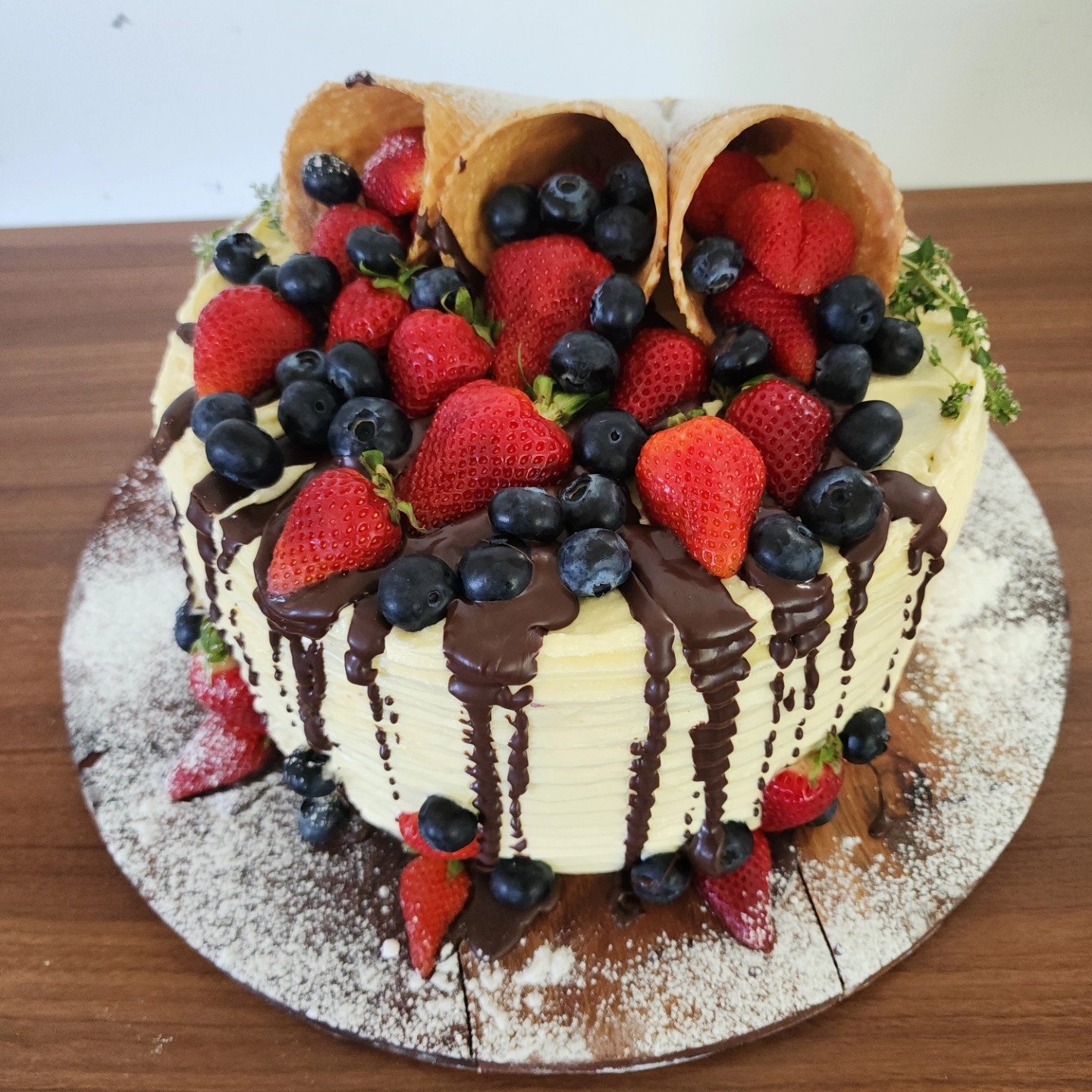 A round cake decorated with fresh strawberries, blueberries, chocolate drizzles, and cannoli shells on top, with powdered sugar on a wooden surface.