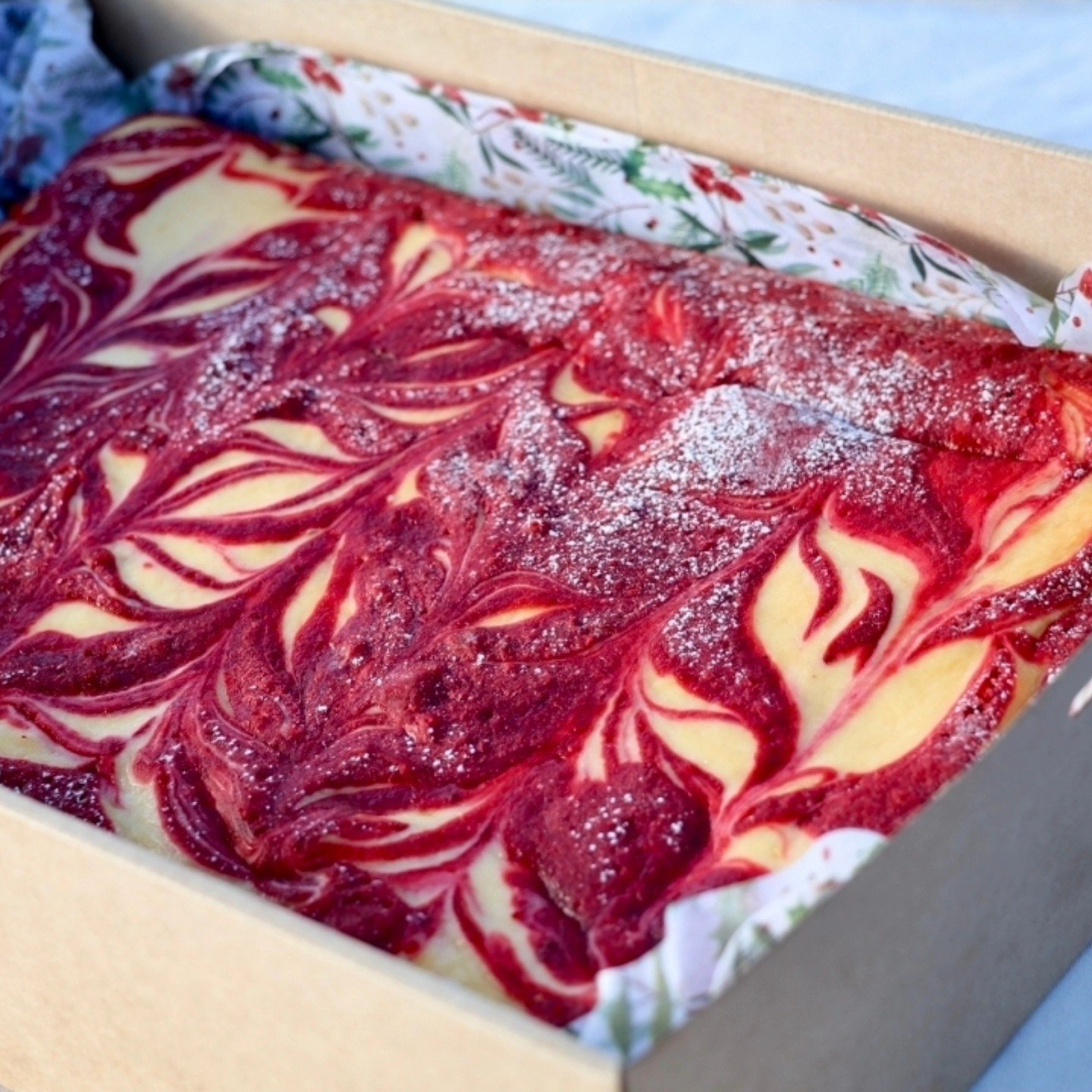 Marbled cheesecake with raspberry swirl in a cardboard box lined with festive holiday paper.