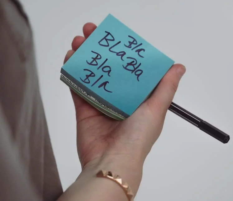 Hand holding a sticky note covered with repetitive writing, representing self-criticism and mental chatter.