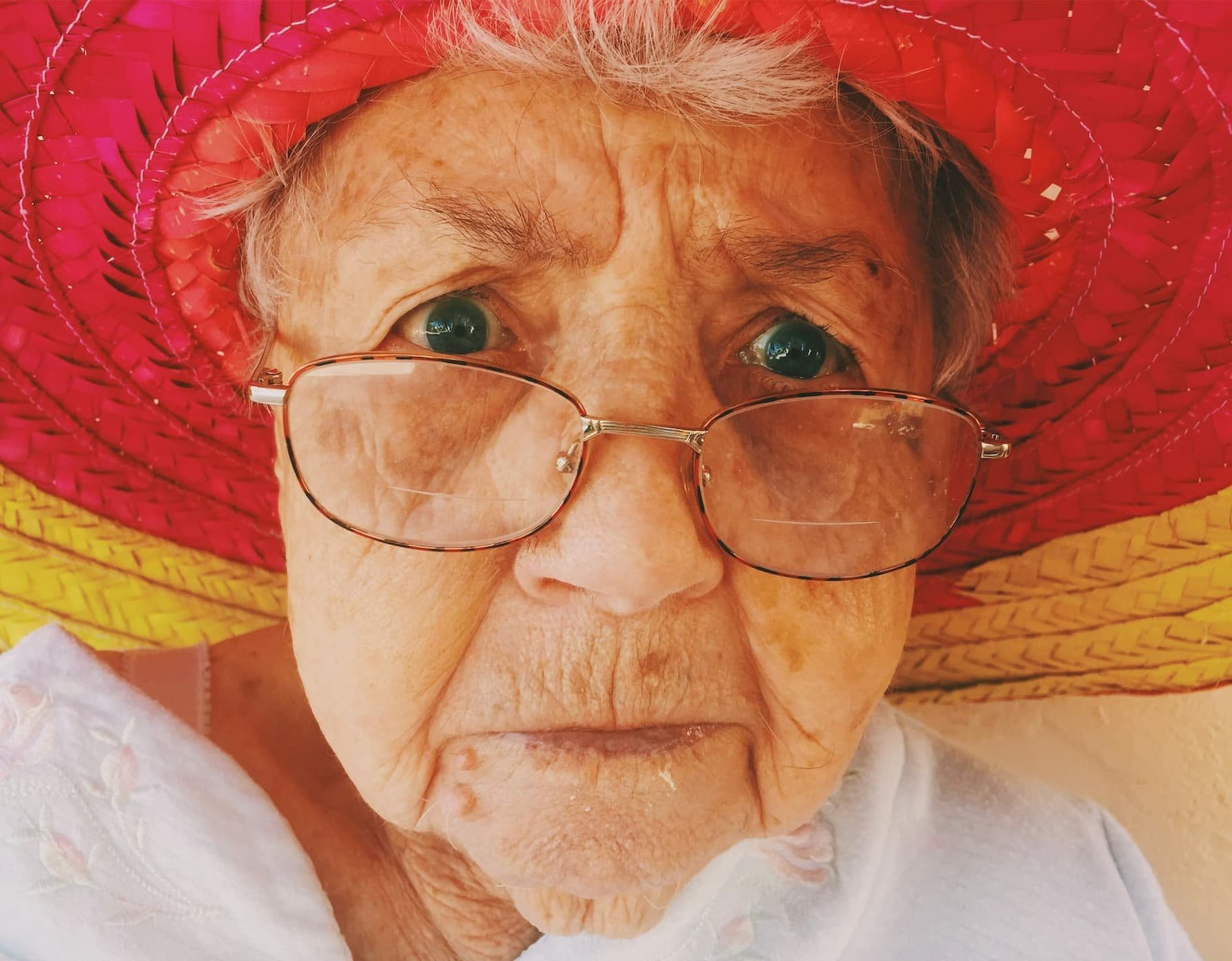 Close-up portrait of an older woman wearing glasses and a bright red hat with a worried, anxious facial expression.