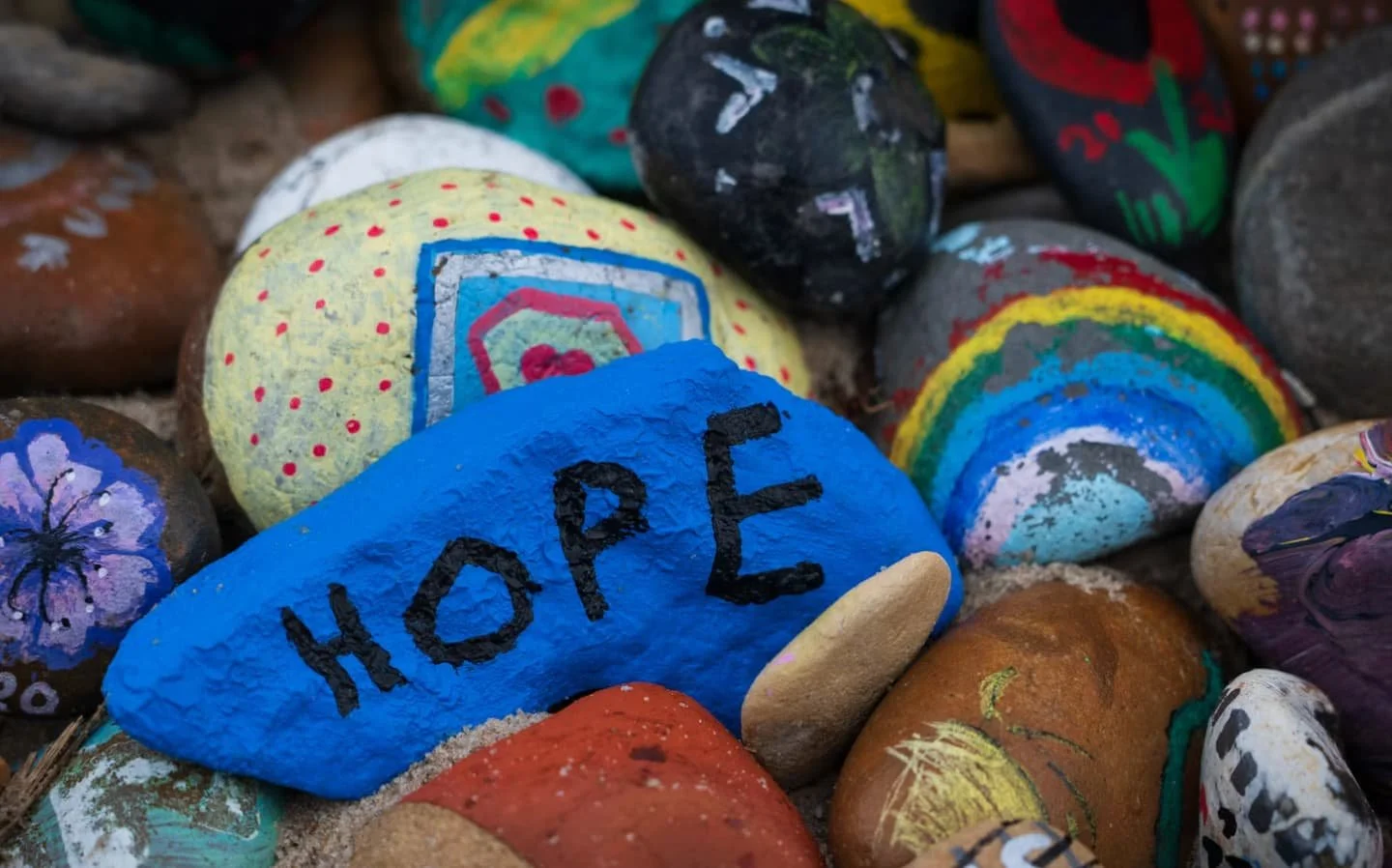 A bright blue painted rock displaying the word "HOPE" among a collection of colorful hand-painted rocks.