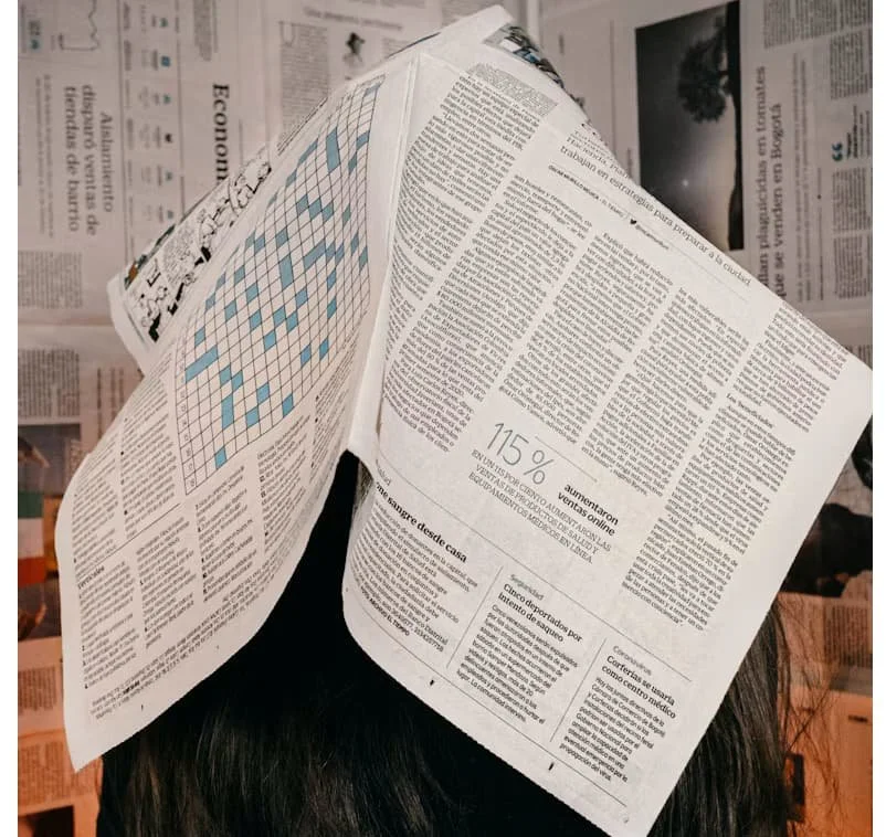Person hiding their face beneath newspapers, surrounded by pages of newsprint