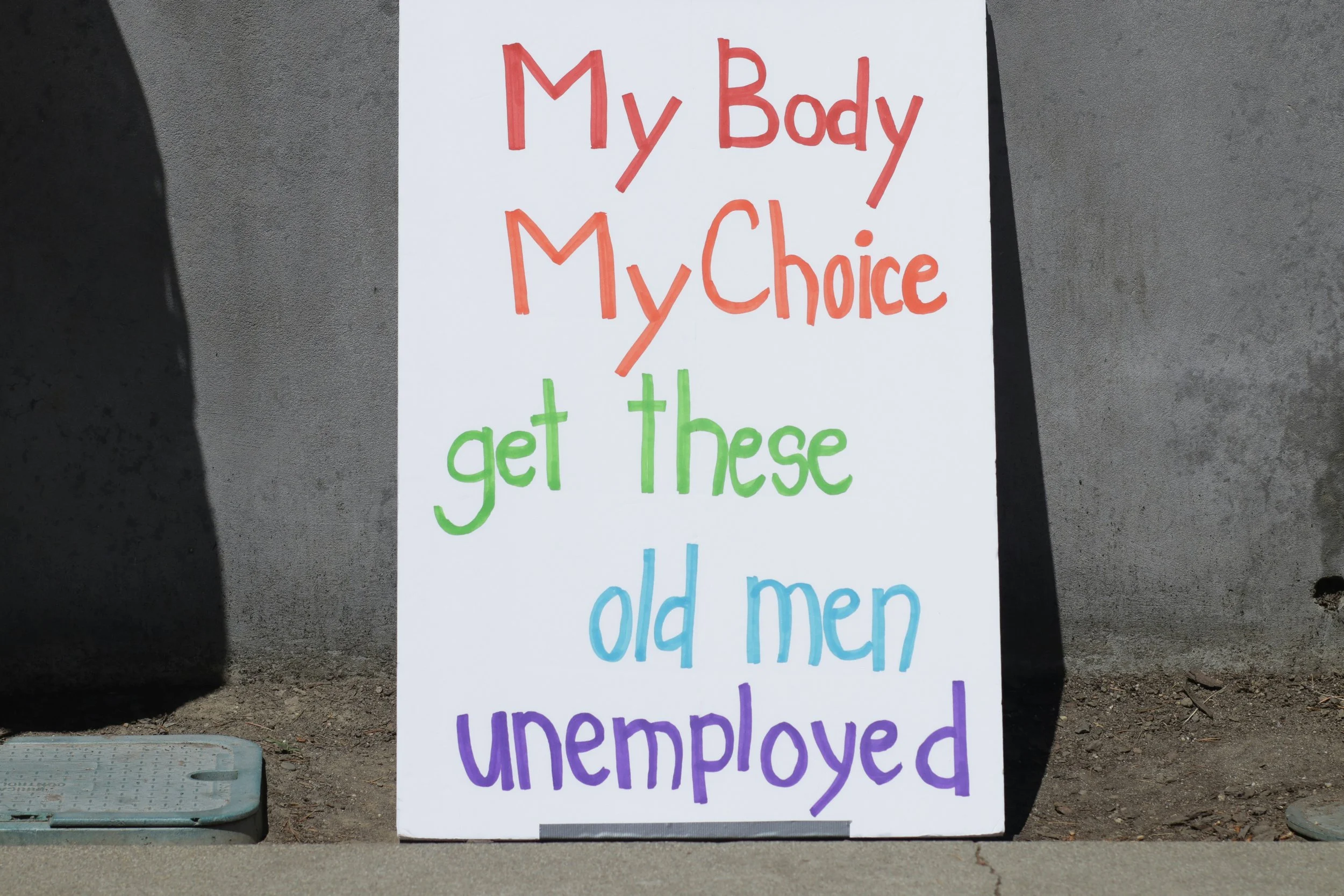A white protest sign with colorful handwritten text reading “My Body, My Choice—get these old men unemployed,” displayed outdoors during a reproductive rights and bodily autonomy demonstration.