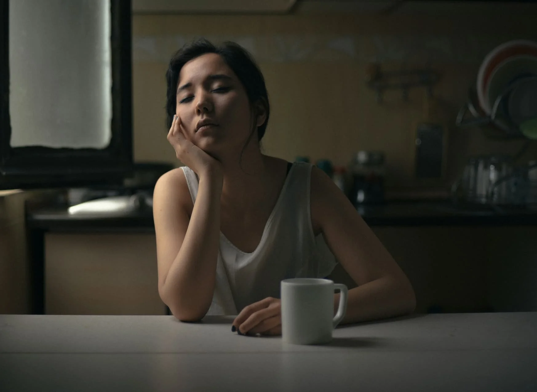 Tired professional woman sitting alone at a kitchen table, deep in thought at the end of the day.