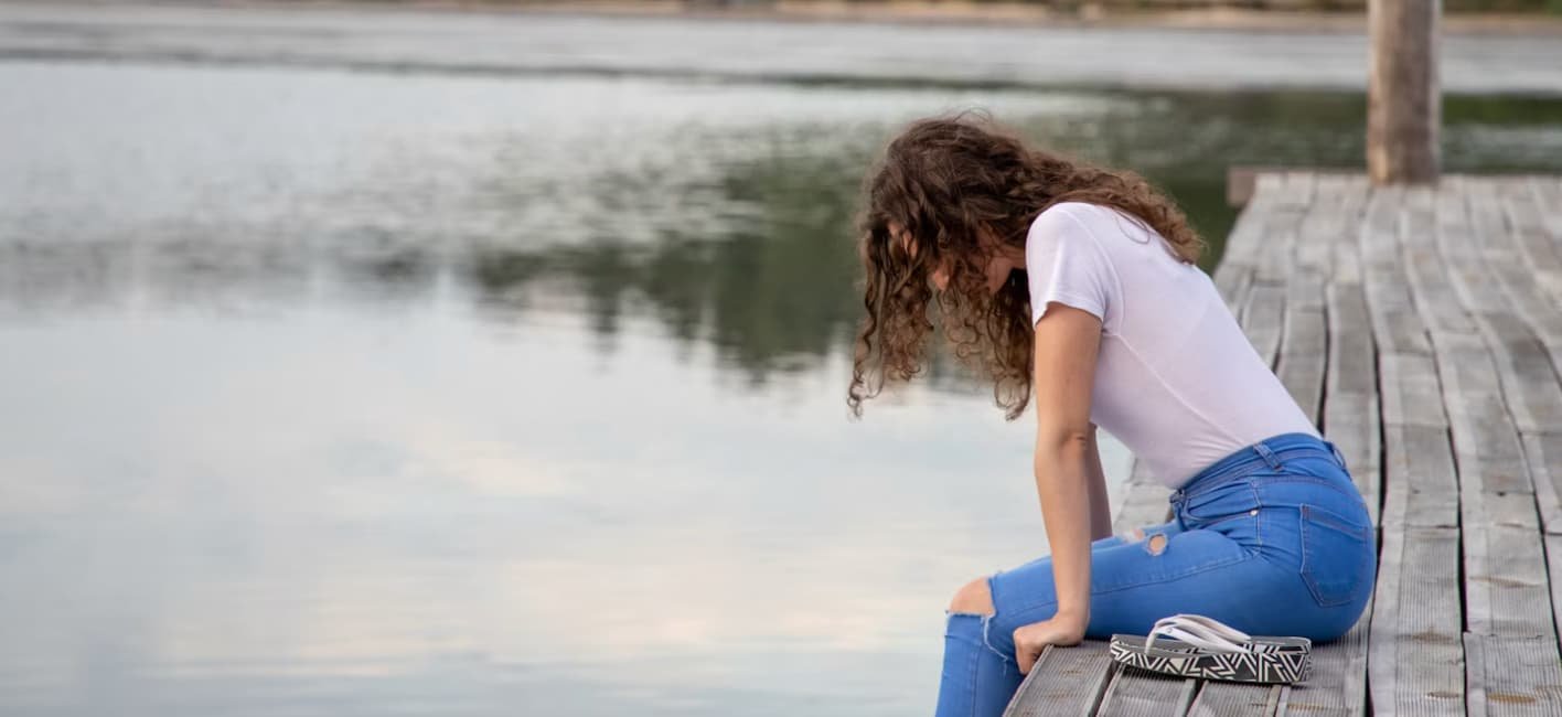 Young woman sitting on a wooden dock with her head bowed, looking out over a calm lake.