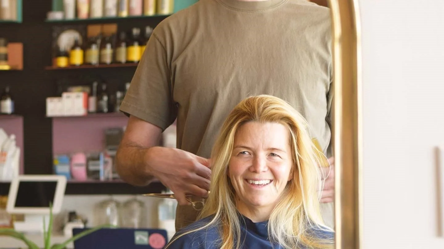 A woman with blonde hair smiling as she gets a haircut in a salon with shelves of products in the background.