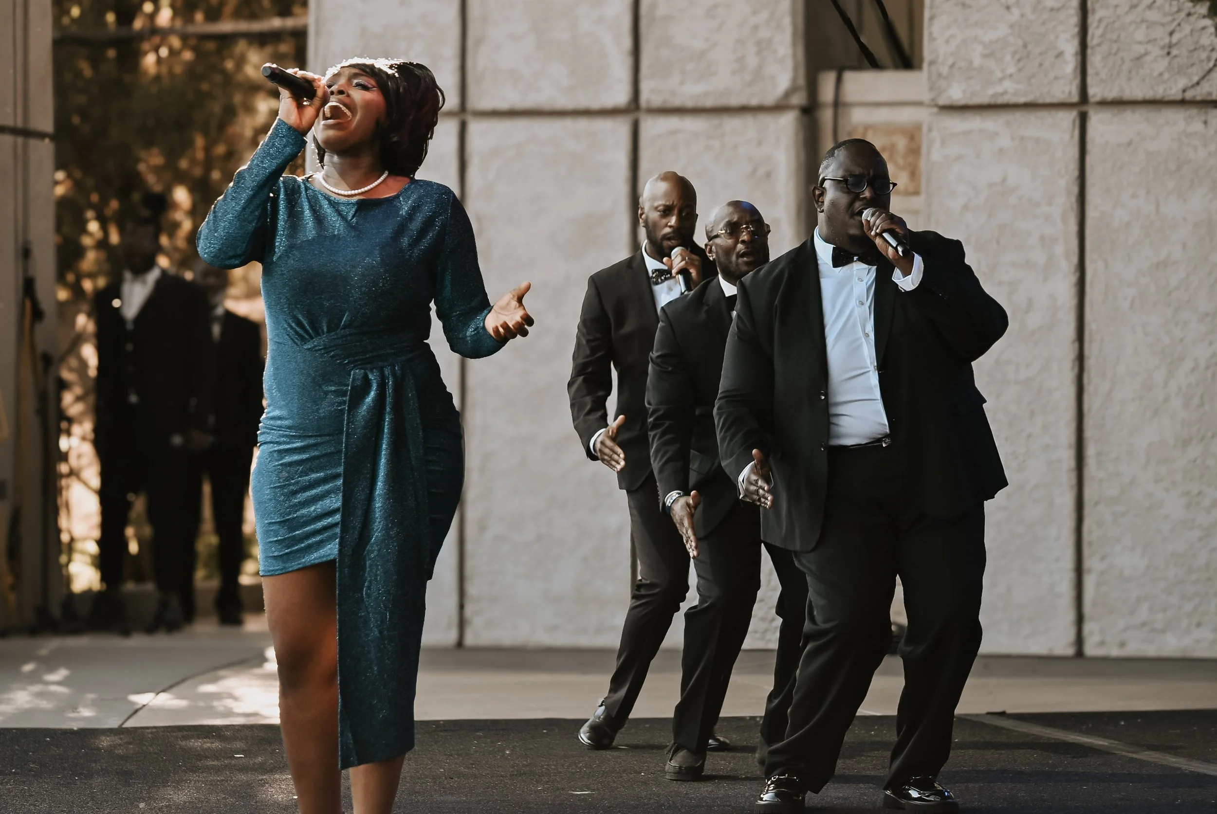 A woman singing with a microphone, wearing a blue dress, with four men in tuxedos singing backup behind her.