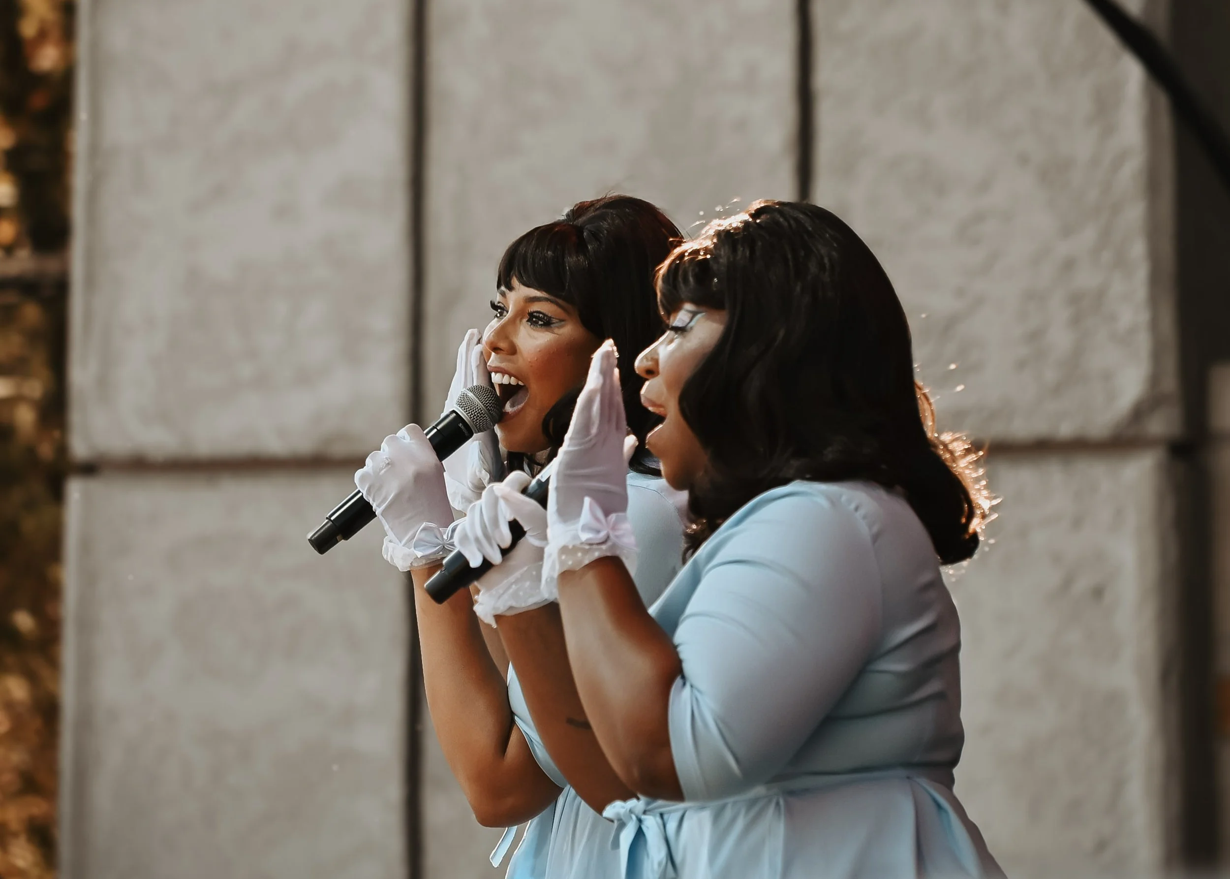 Two women dressed as maids, both holding microphones and wearing white gloves, appear to be speaking or singing. They are in an indoor setting with a plain, light-colored wall in the background.