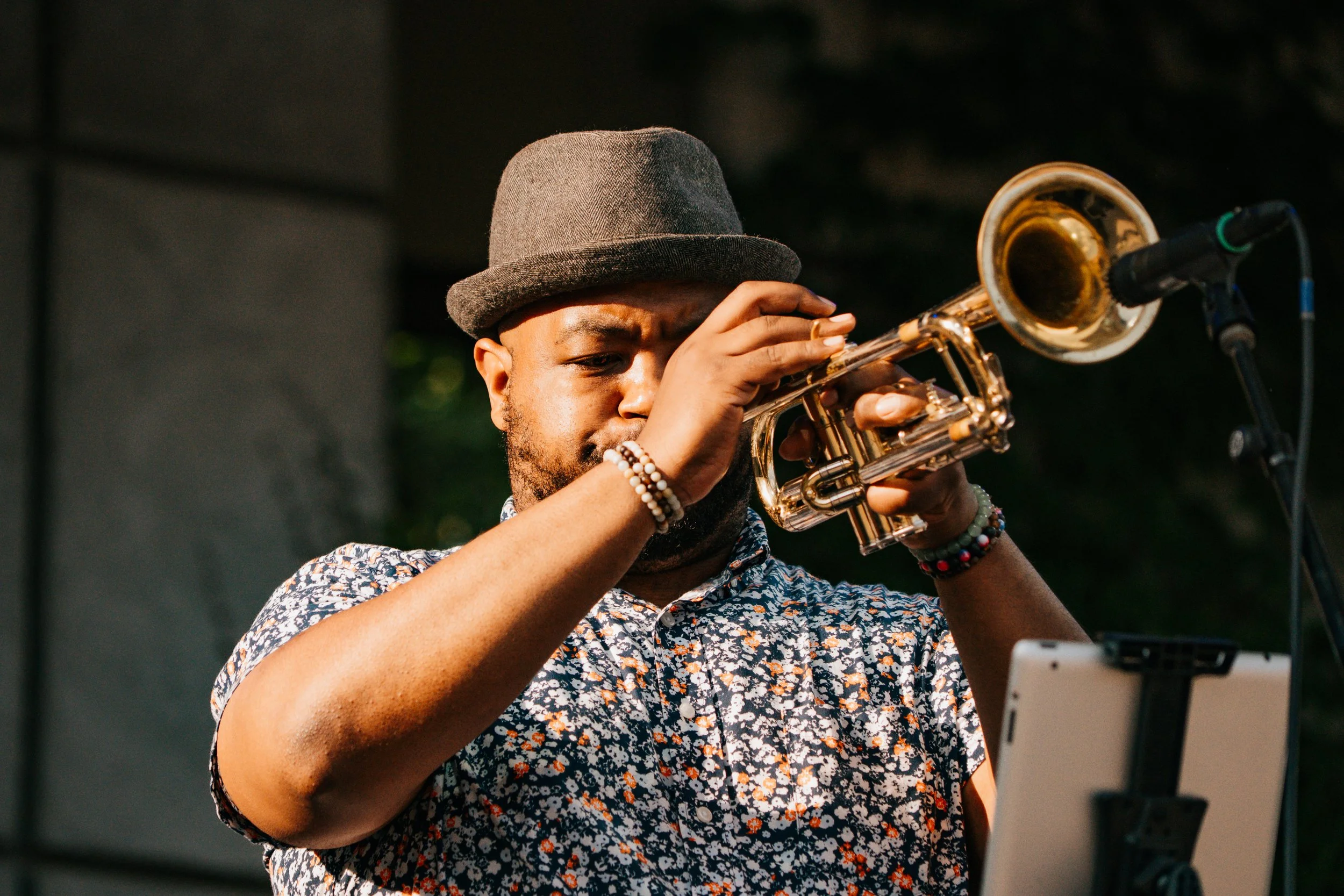 A man wearing a gray hat and a floral shirt, playing a trumpet on stage.