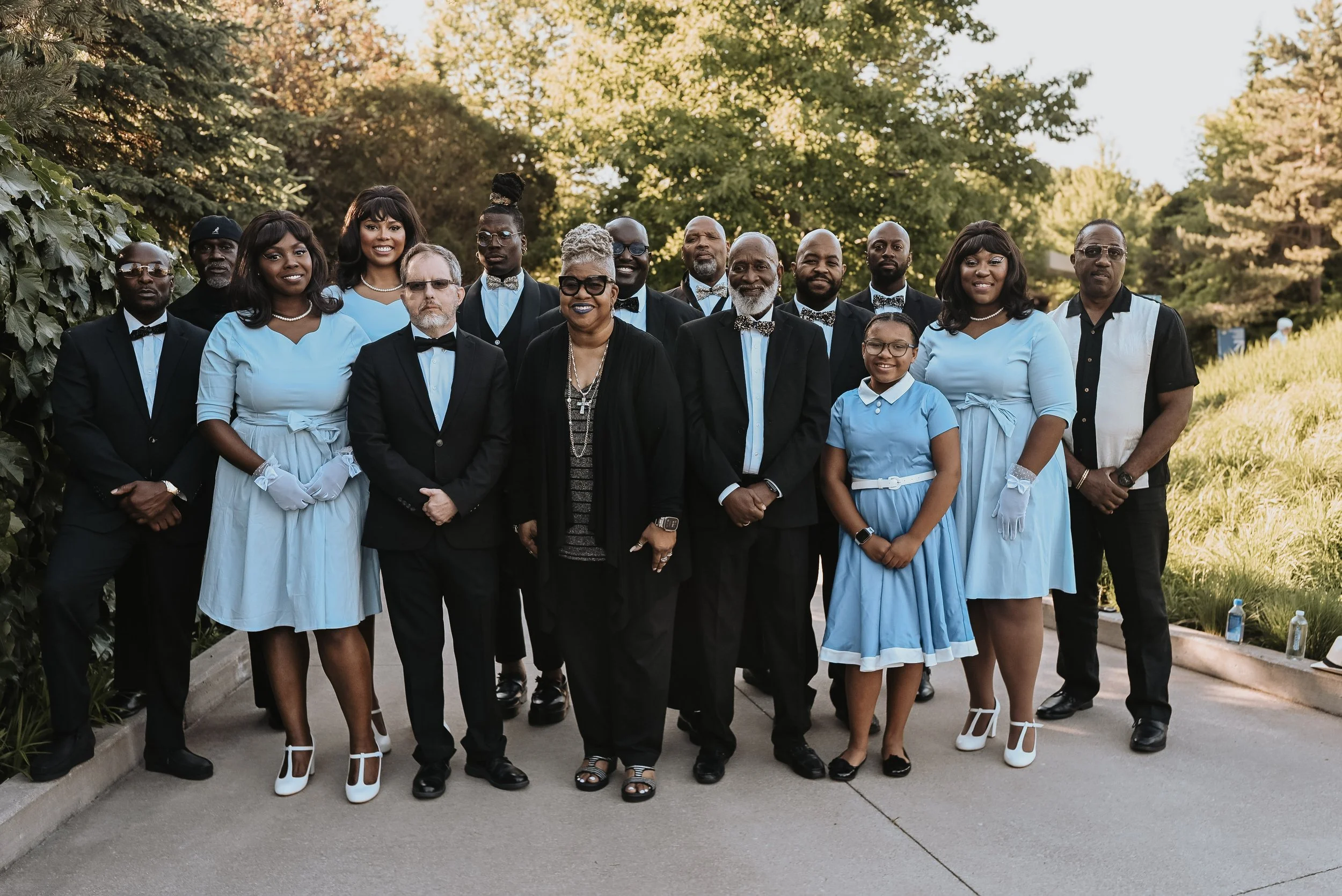 Group of 16 people posing outdoors for a photo, dressed in formal attire. The women are wearing light blue dresses and white gloves, while the men are in suits with bow ties. Green trees and foliage are in the background.