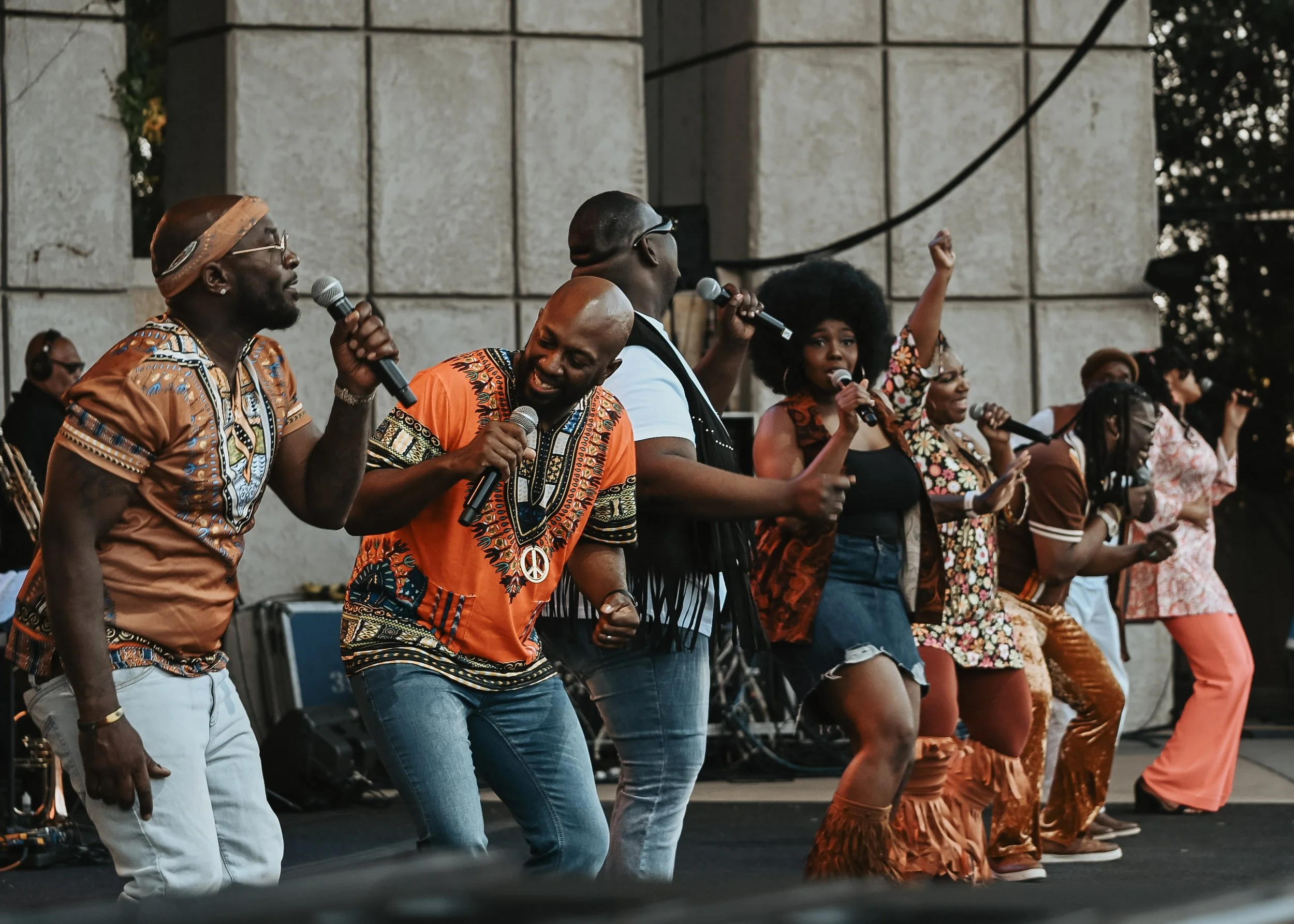 A group of people dancing and singing on an outdoor stage, dressed in colorful clothing with African patterns.