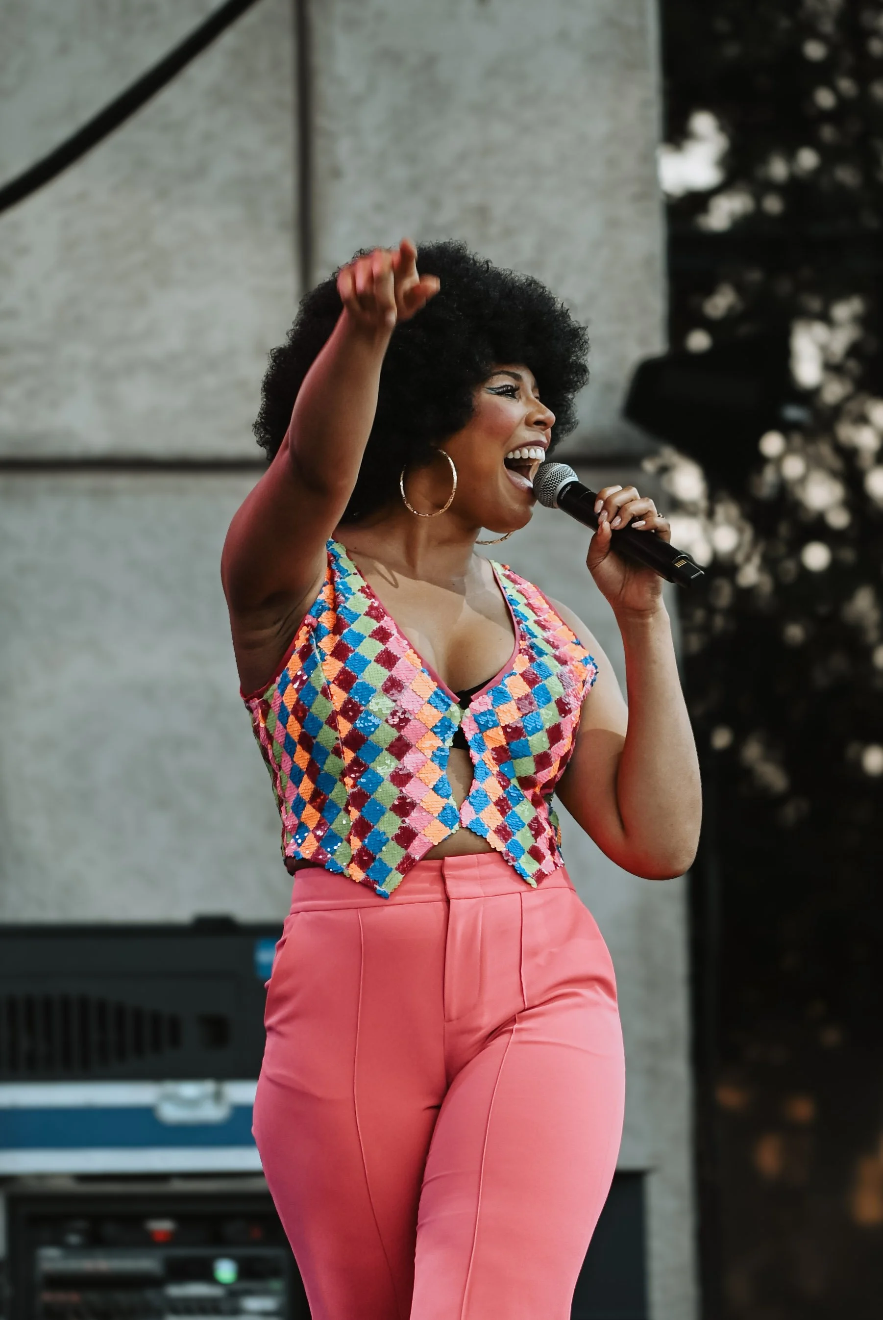 A woman with curly hair, large hoop earrings, wearing a colorful checkered crop top and pink high-waisted pants, singing into a microphone and raising her right hand with a joyful expression.