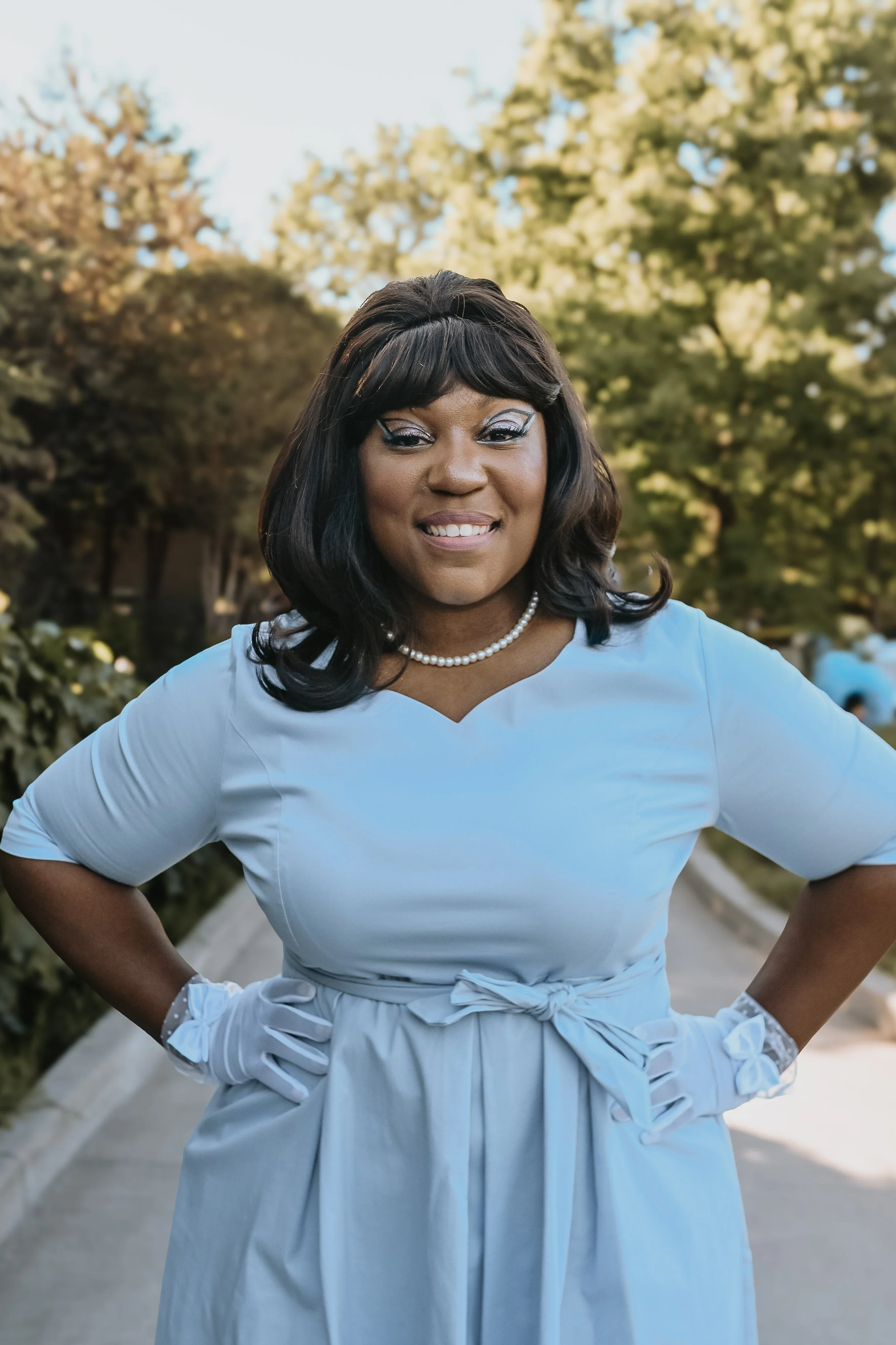 A woman smiling outdoors in a light blue dress with a pearl necklace, standing on a pathway with trees in the background.