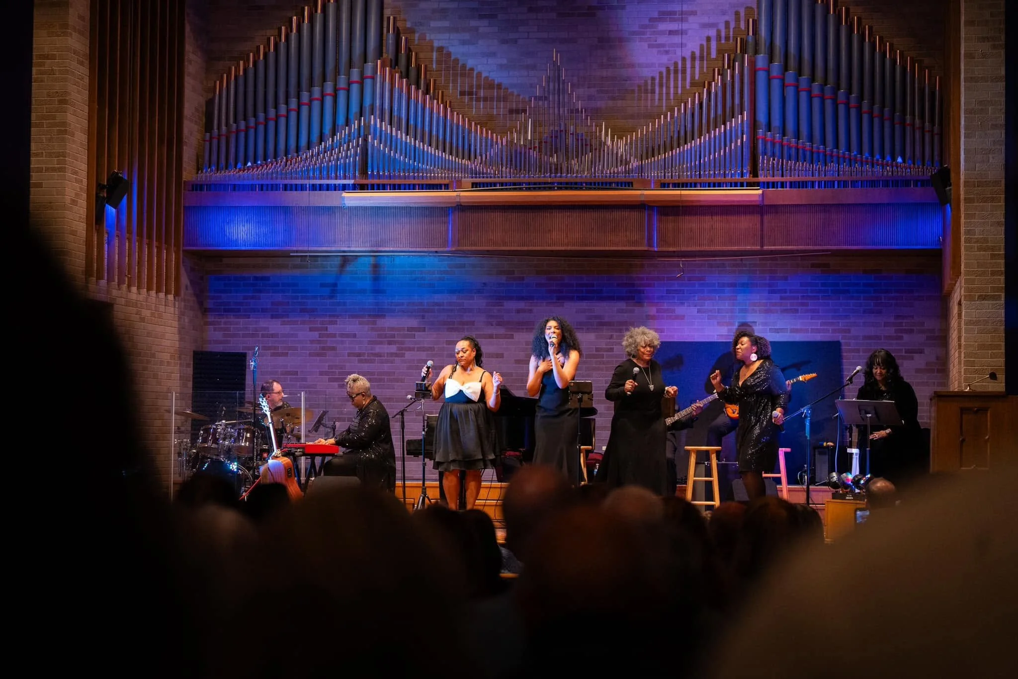 A group of musicians and singers performing on stage with instruments including drums, keyboard, and guitar, in front of a large pipe organ and brick wall background, illuminated with purple and blue stage lighting.