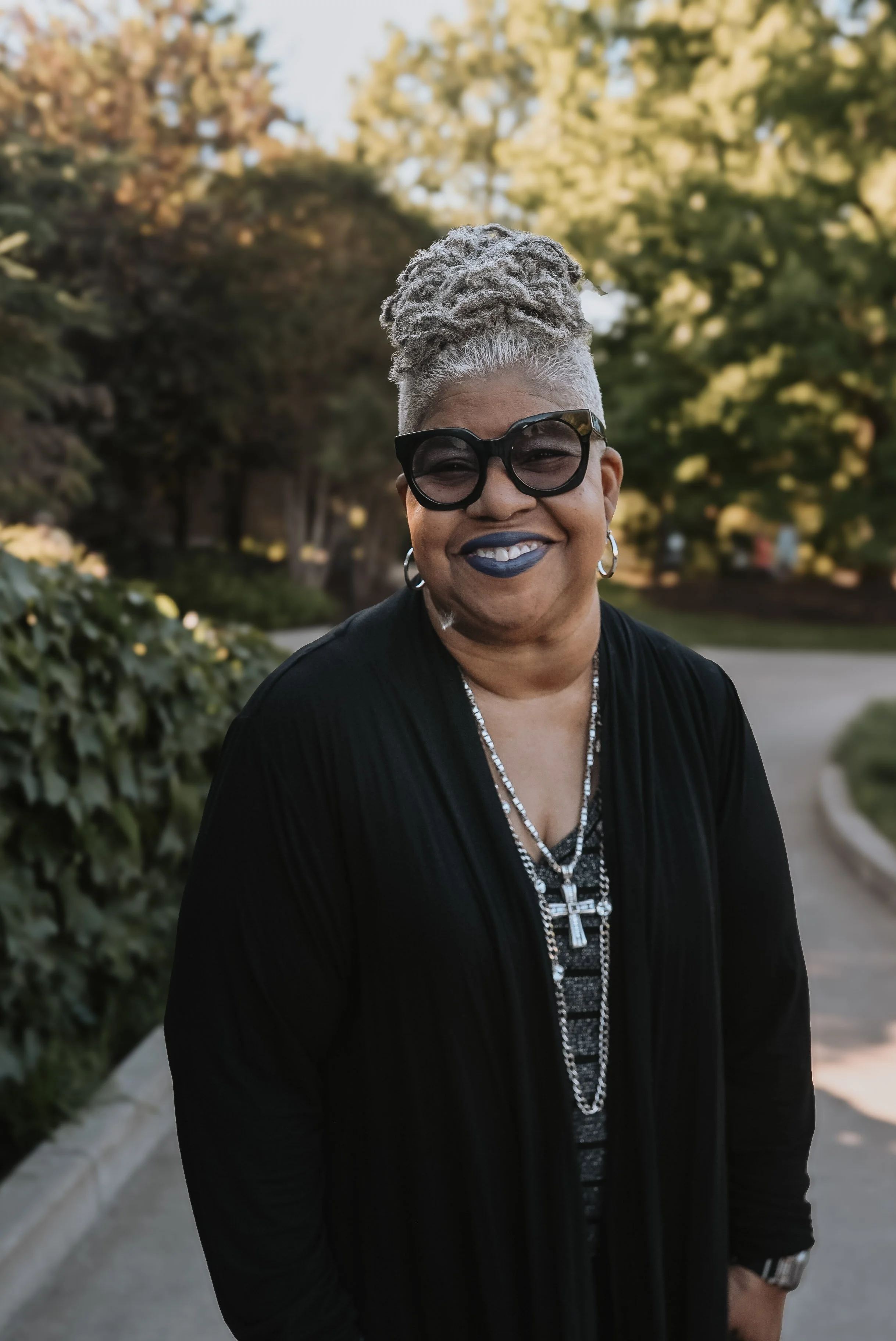A smiling woman with gray dreadlocks in an updo, black sunglasses, dark lipstick, hoop earrings, wearing a black jacket with patterned shirt underneath, standing outdoors during daylight with trees and a sidewalk in the background.