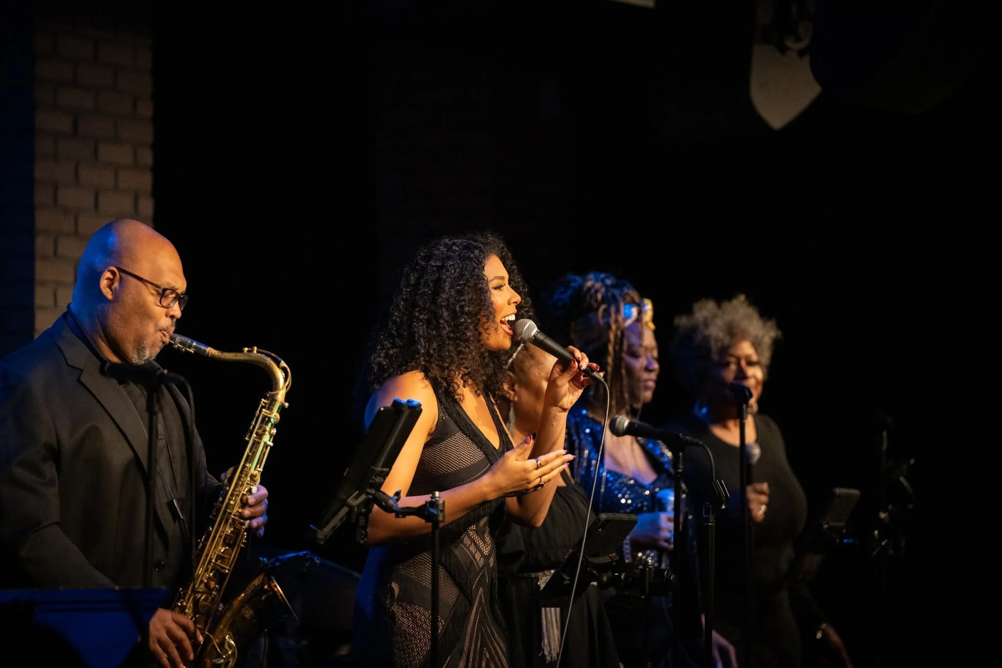 A group of musicians performing on stage, including a woman singing into a microphone and a man playing the saxophone, with others in the background.