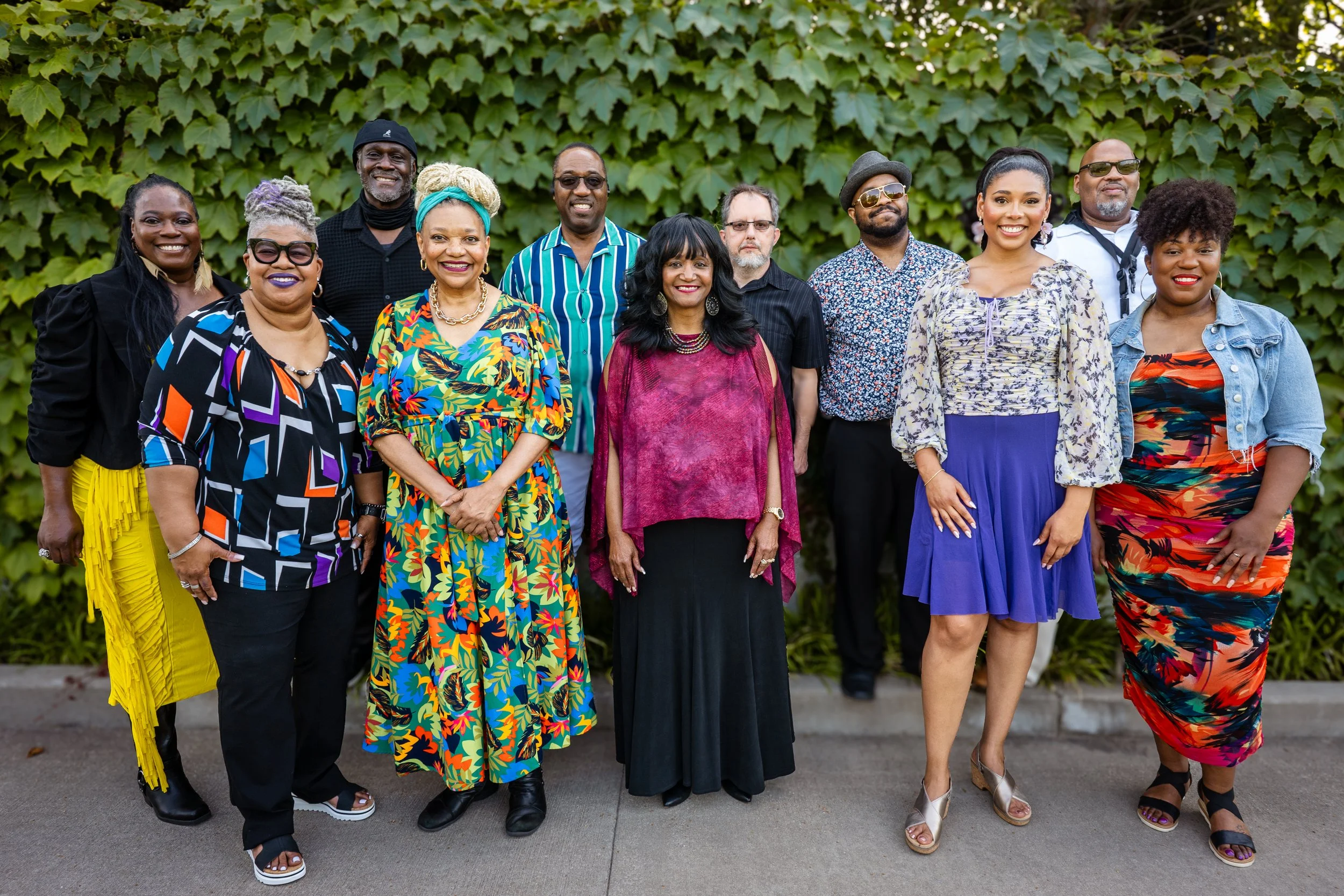 Group of diverse people standing outdoors in front of green leafy background, smiling, dressed in colorful and stylish clothing.