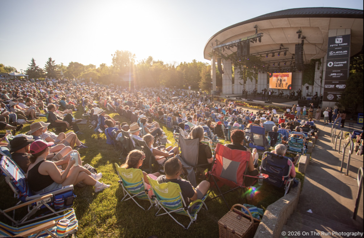 Large outdoor concert with a crowd seated on the grass and in chairs, sun setting behind trees, stage with performers and a large screen displaying 'Congratulations'.