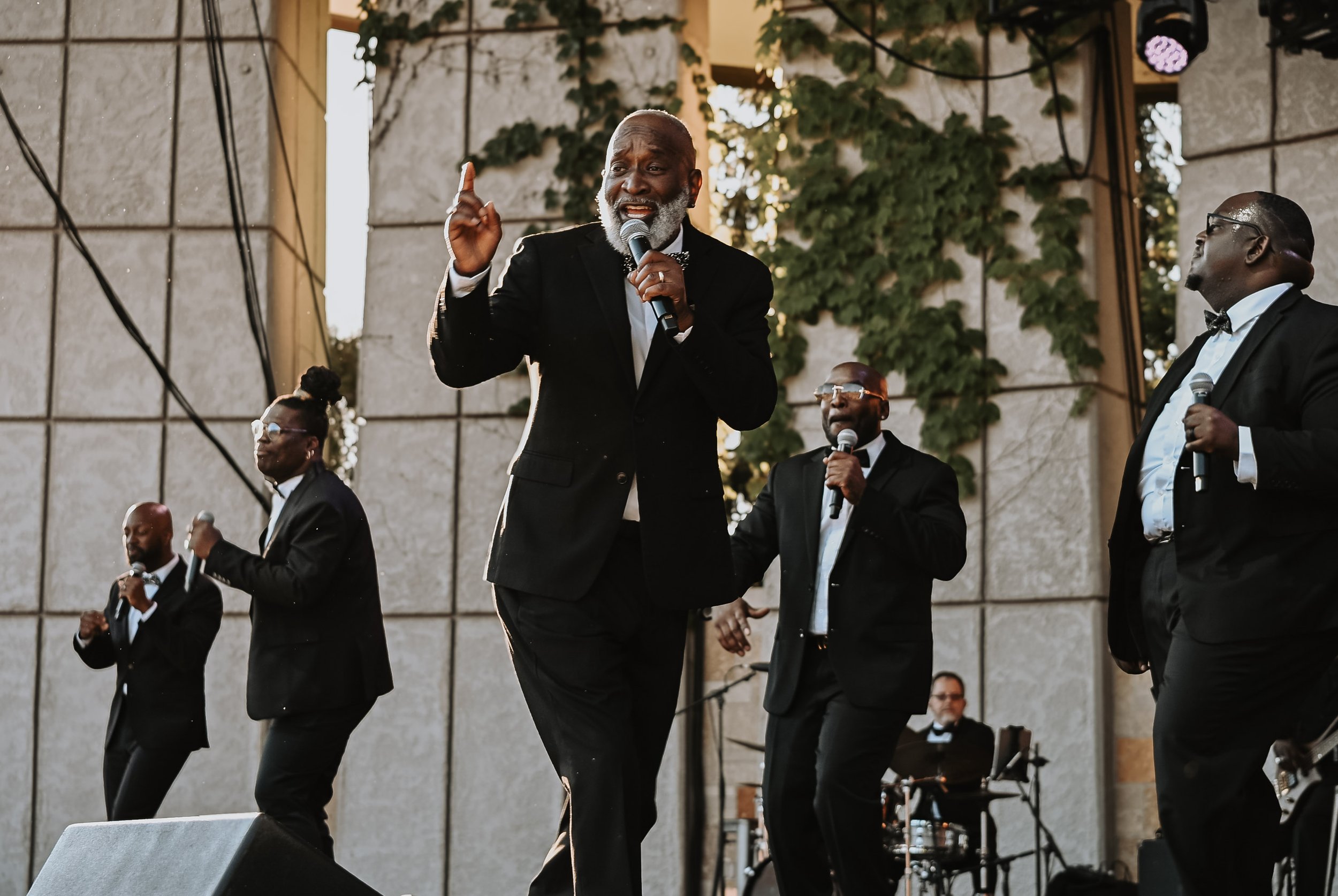 A group of people dressed in formal attire performing on an outdoor stage, with one man singing into a microphone while others hold microphones and sing. The stage has a backdrop of greenery and a stone wall.