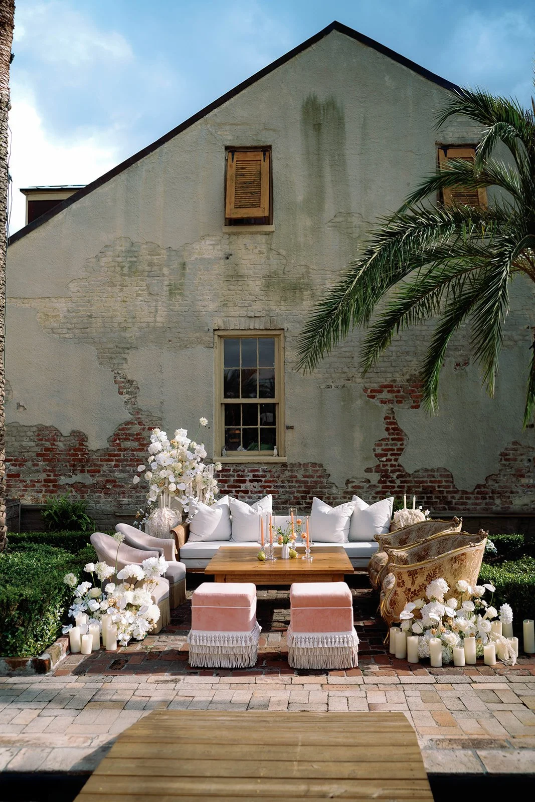 Outdoor patio decorated for a celebration with a white sofa, vintage armchairs, a wooden table, pink ottomans, white flowers, candles, and greenery against an old brick wall with peeling paint and a window, under a blue sky.